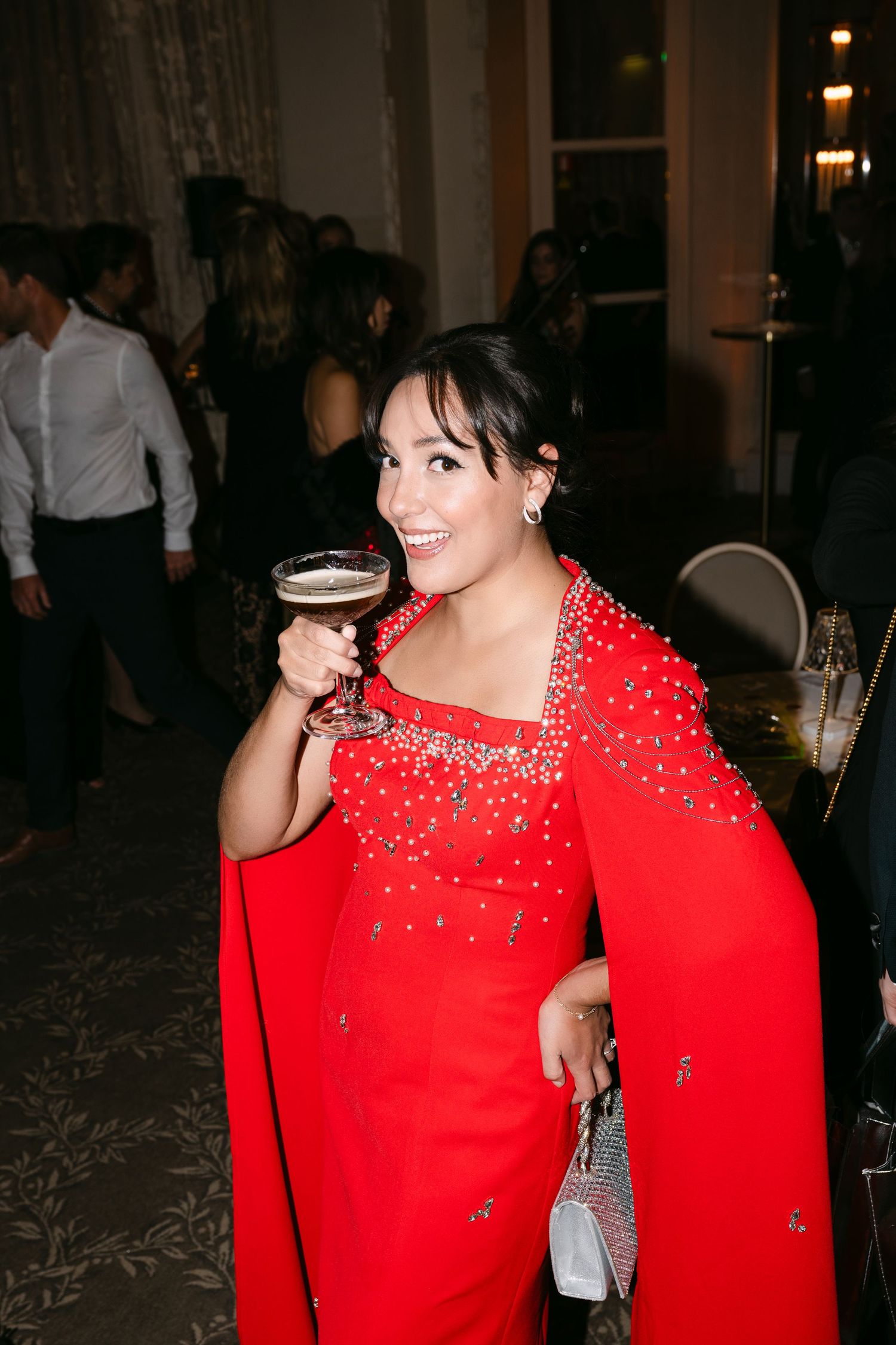 Smiling woman in red embellished cape gown enjoys espresso martini cocktail at elegant wedding reception party.