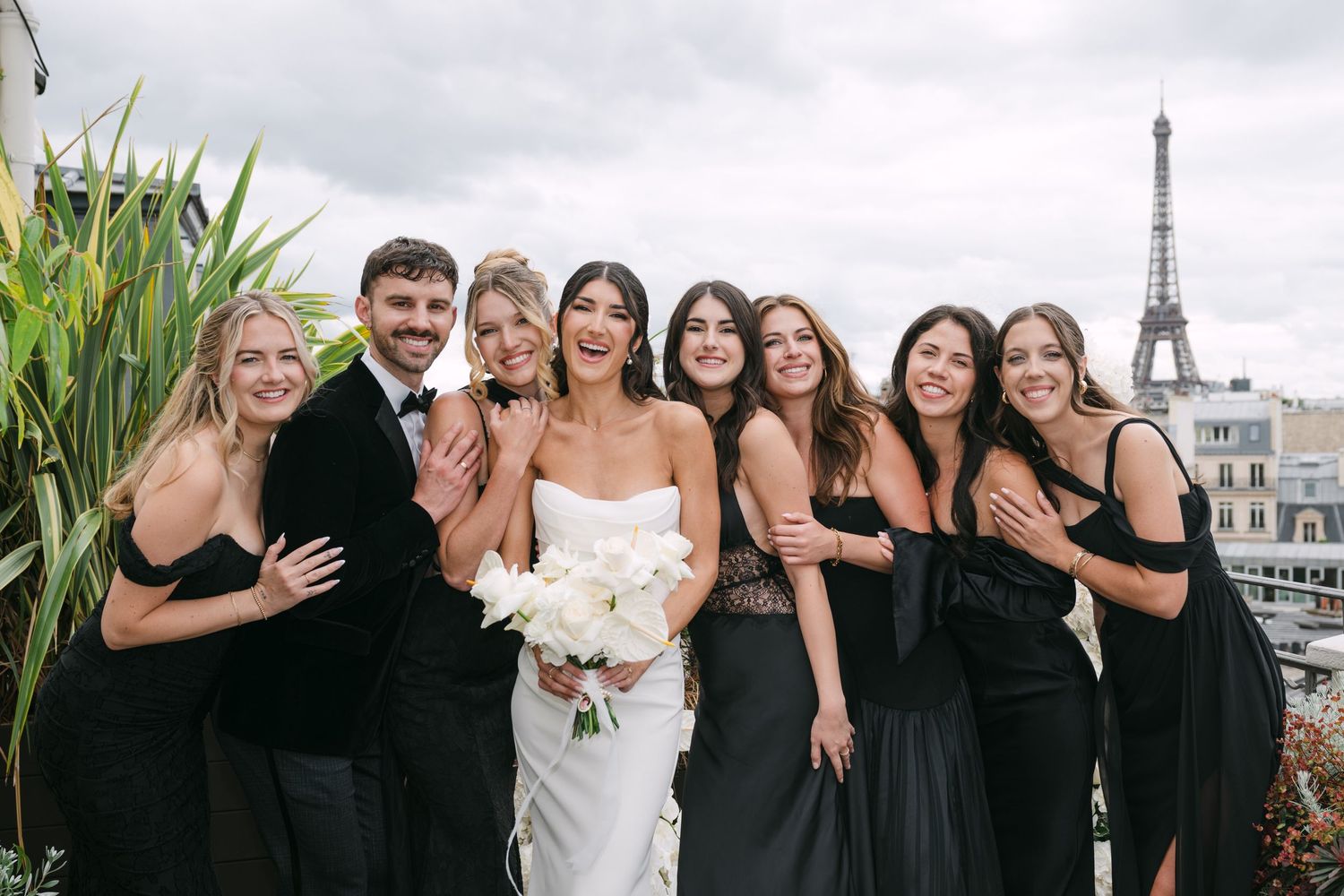 Bride in white gown with bridesmaids in black dresses smiling on Paris rooftop near Eiffel Tower.