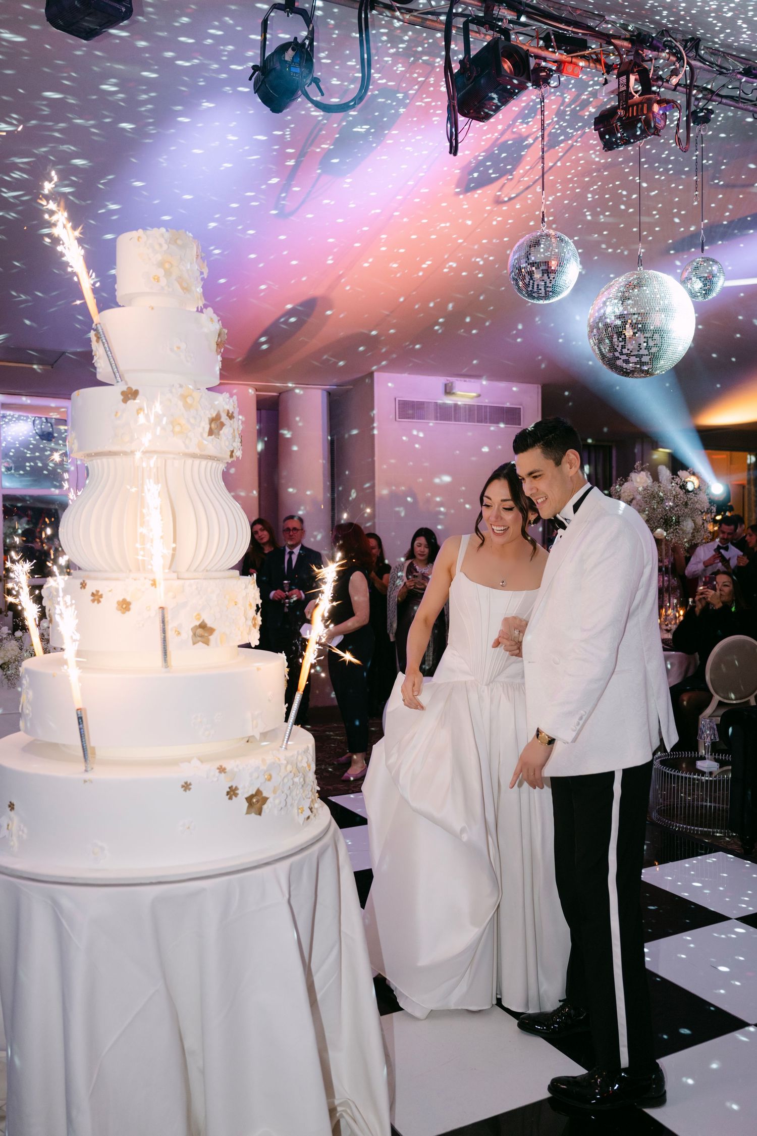 Bride and groom cutting tall white wedding cake with sparklers and confetti on dance floor.