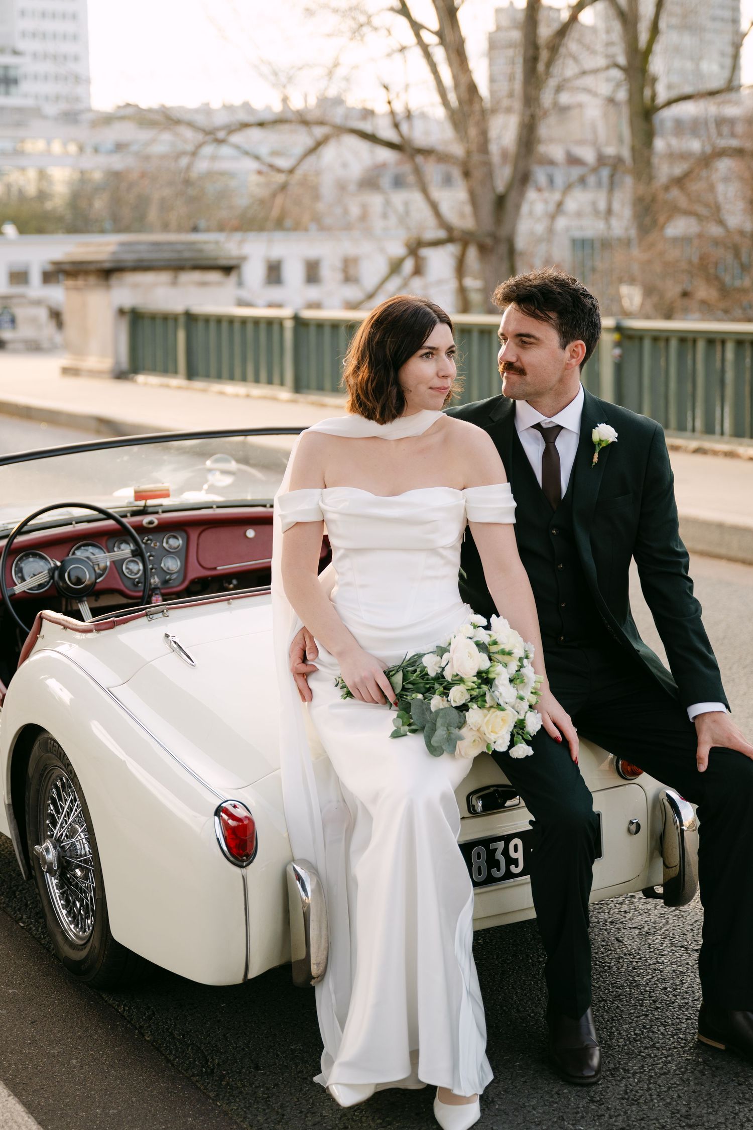 Bride and groom pose beside a vintage white convertible car on a bridge, bride holding white bouquet in wedding attire.