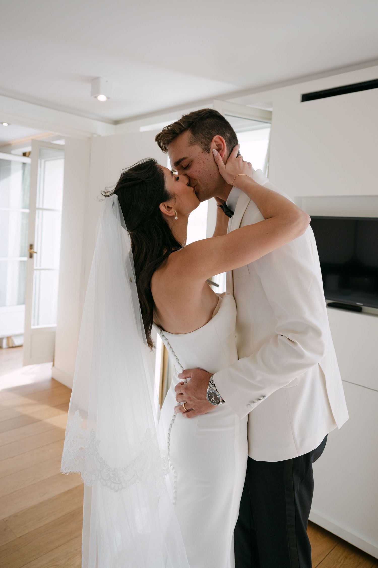 A couple embraces in a bright white room with hardwood floors and natural light streaming through windows.