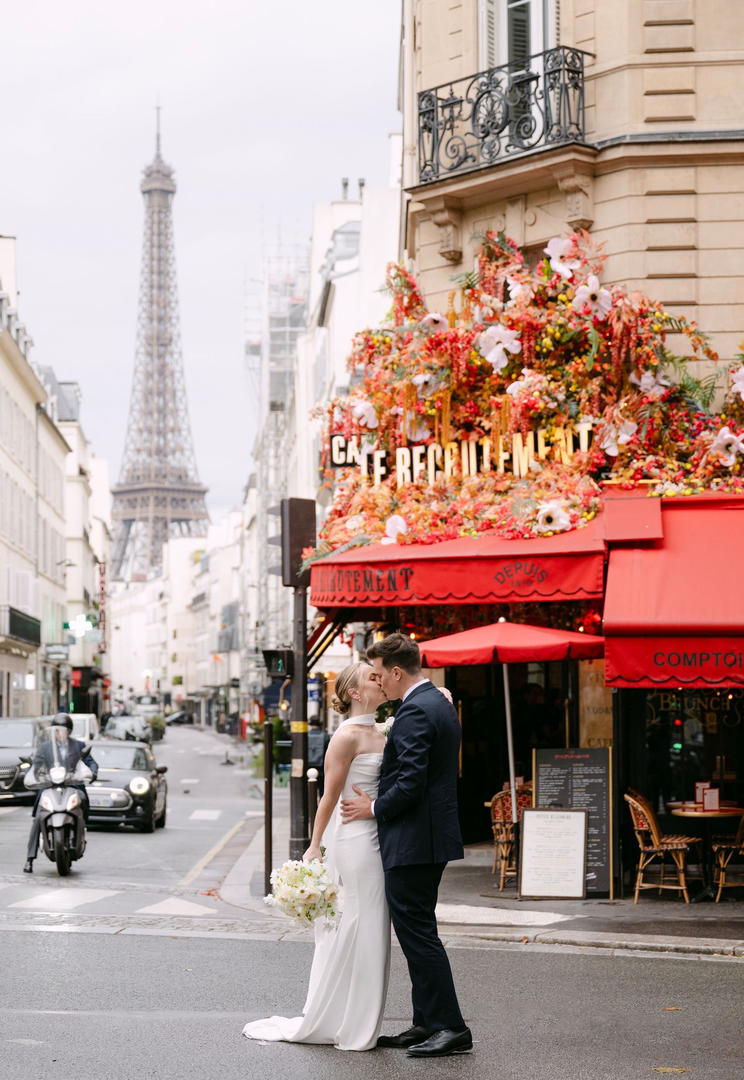 Romantic couple poses outside a charming Parisian cafe with red awning and the Eiffel Tower visible in the background.