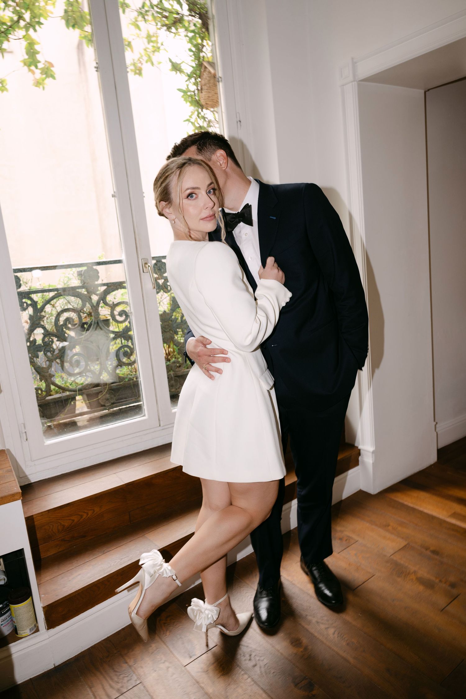 A couple in formal attire poses playfully by a window, with her in a short white dress and him in a black tuxedo.