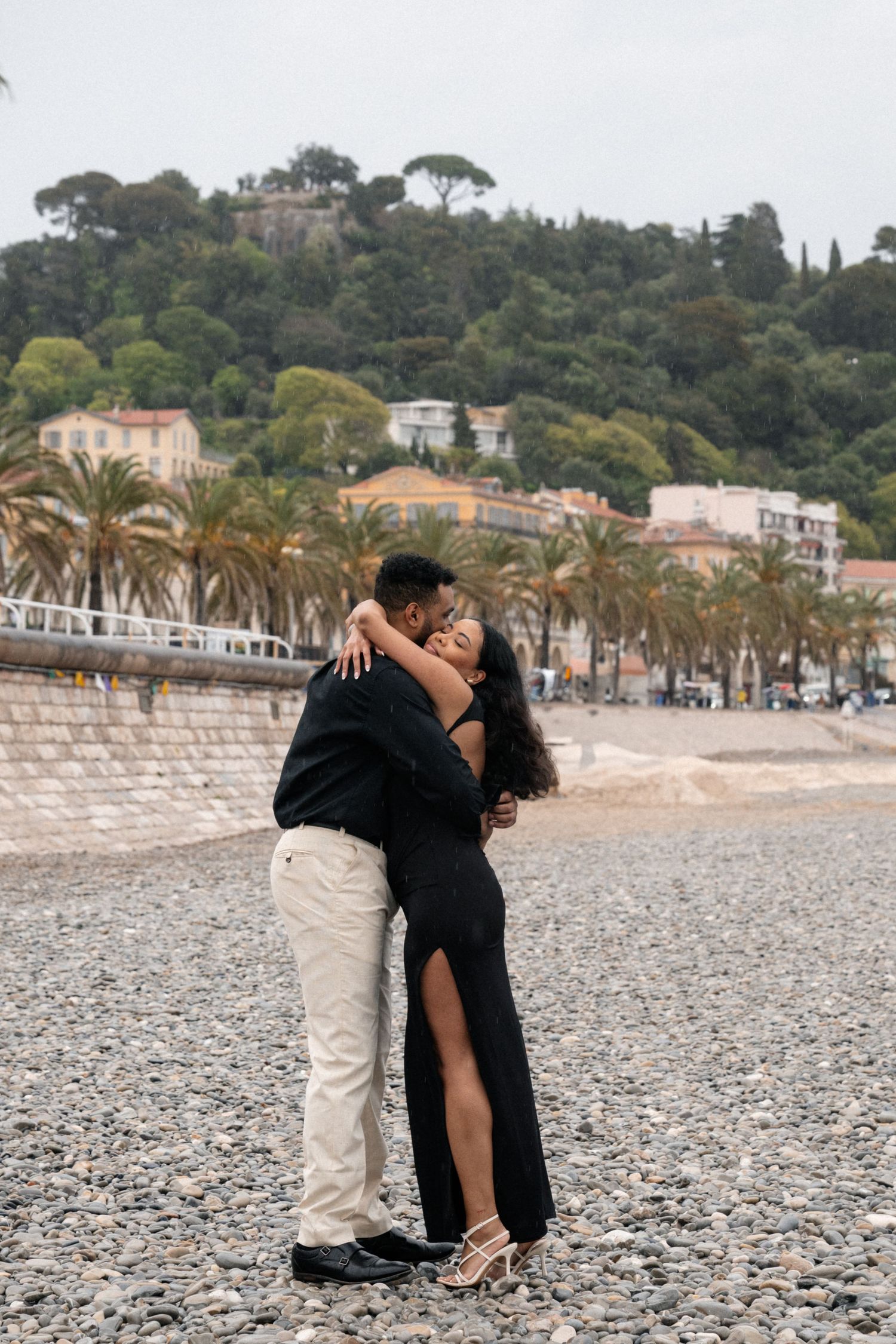 A couple embraces on a pebbly beach with palm trees and hillside buildings in Nice, France.