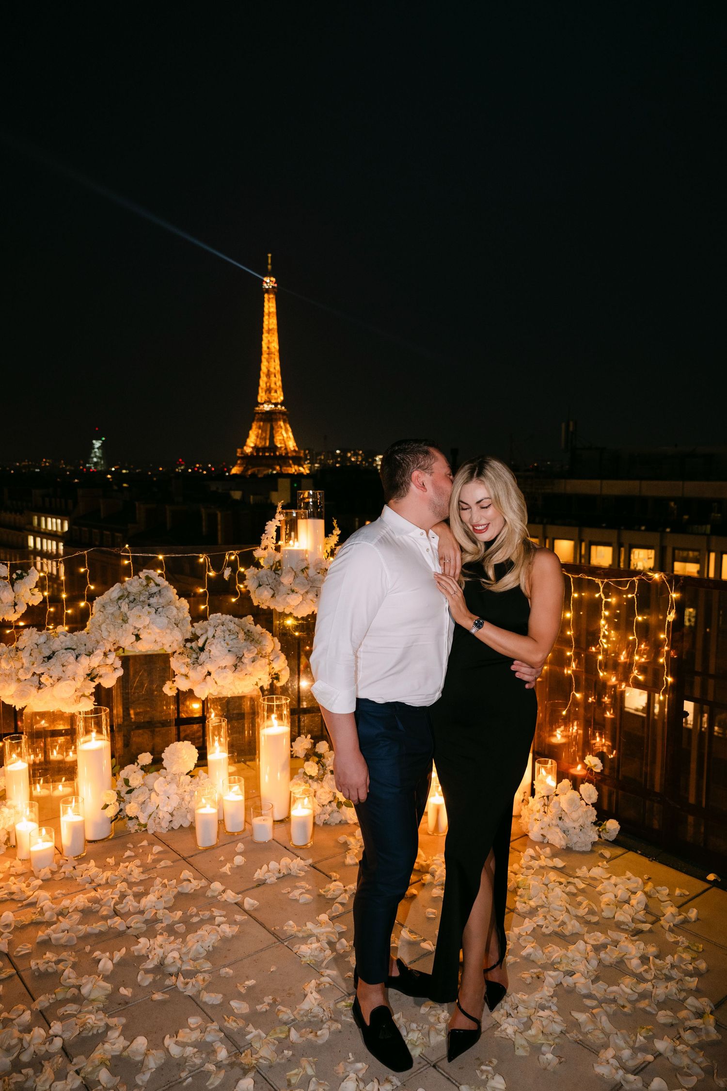 Romantic couple embraces on candlelit rooftop with illuminated Eiffel Tower glowing at night.
