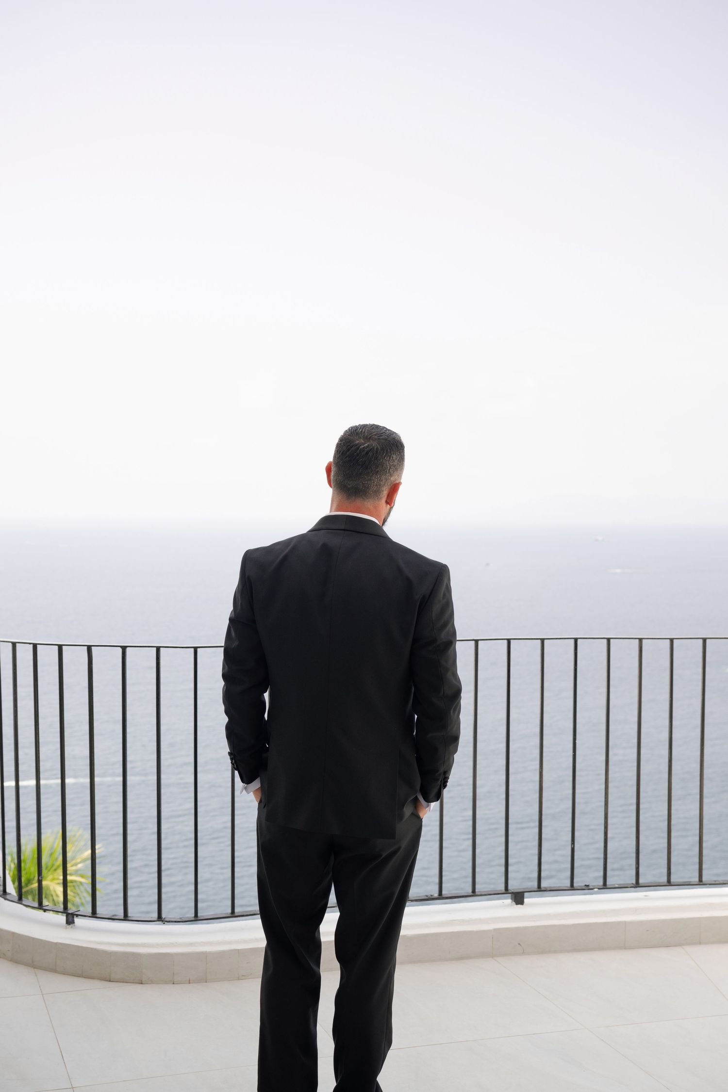 Man in black suit standing on seaside balcony overlooking calm ocean waters.