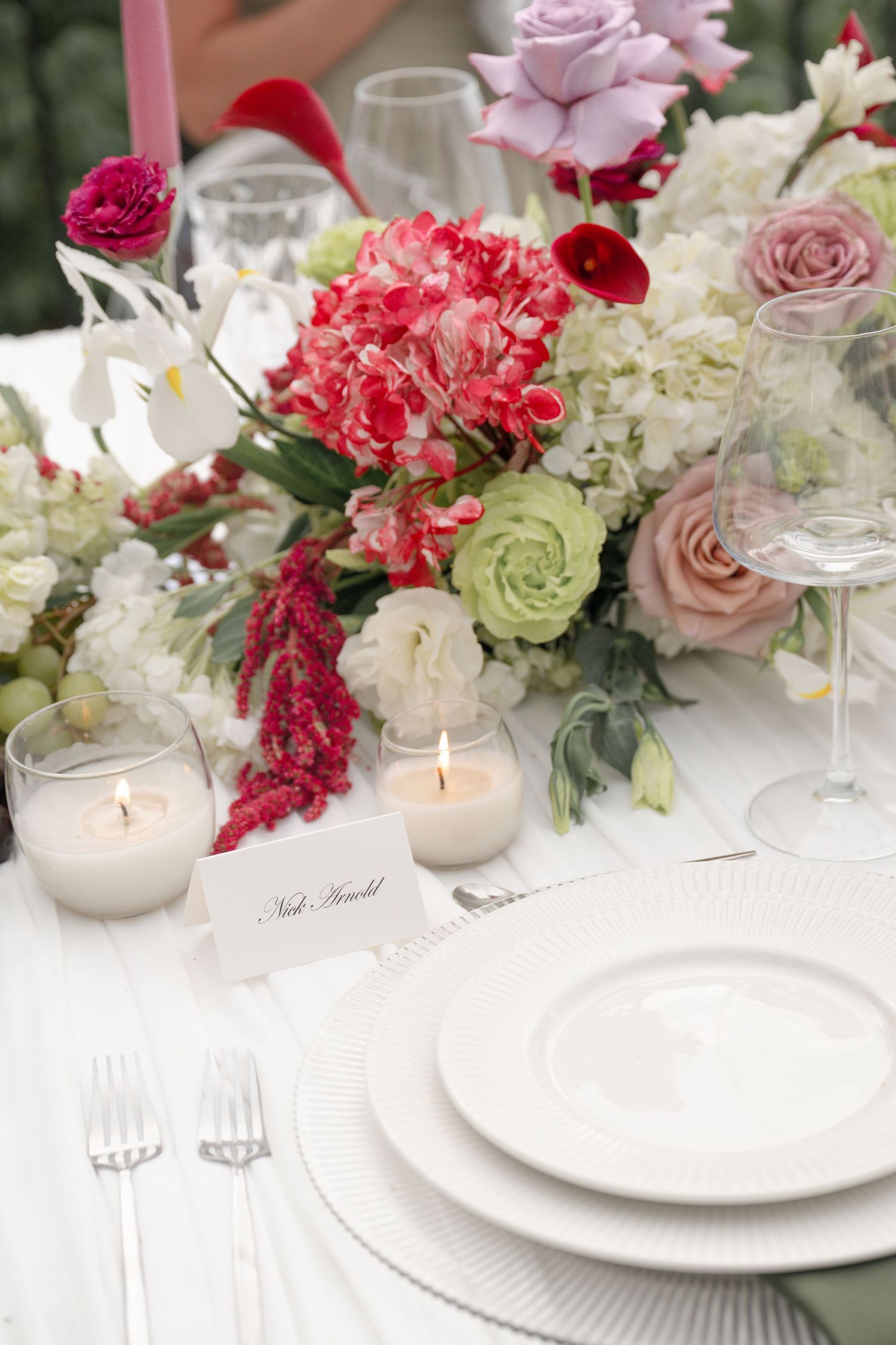 Romantic wedding table setting with pink and red floral arrangement, lit candles, and white place settings.