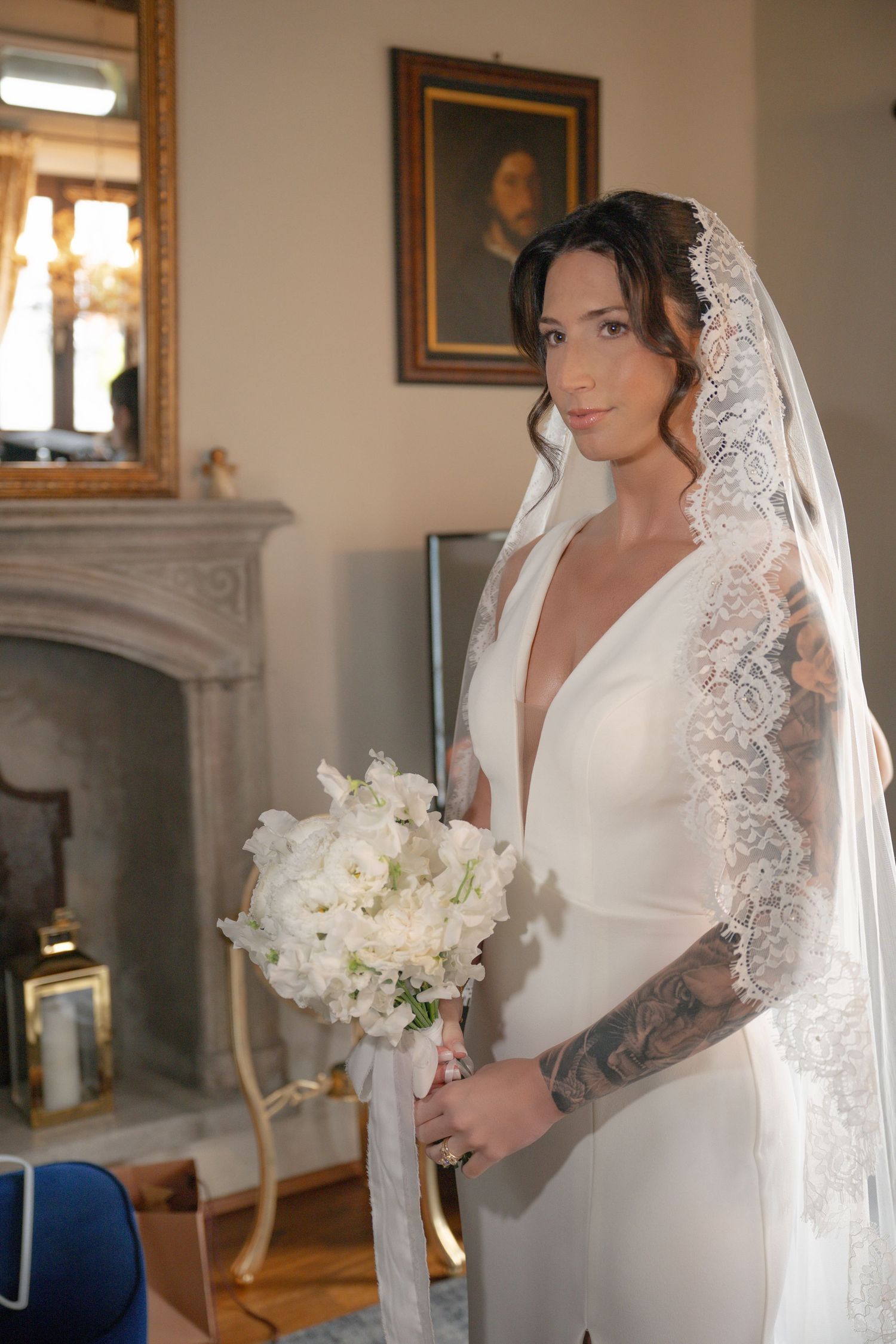 Tattooed bride in deep-V white gown and lace veil holds white bouquet beside fireplace in elegant interior.