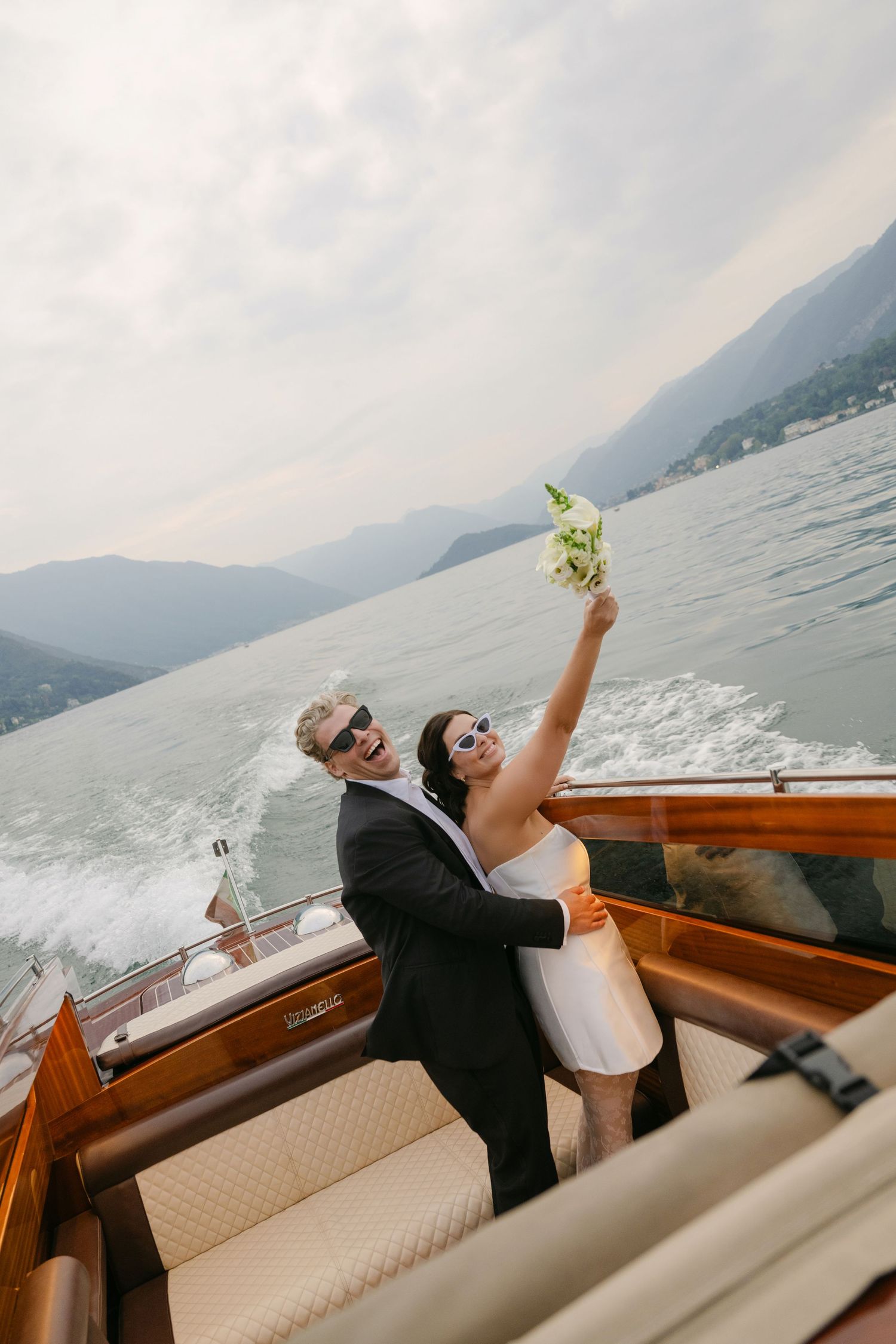Bride and groom celebrate on a wooden boat on Lake Como, Italy, with mountains in the background.