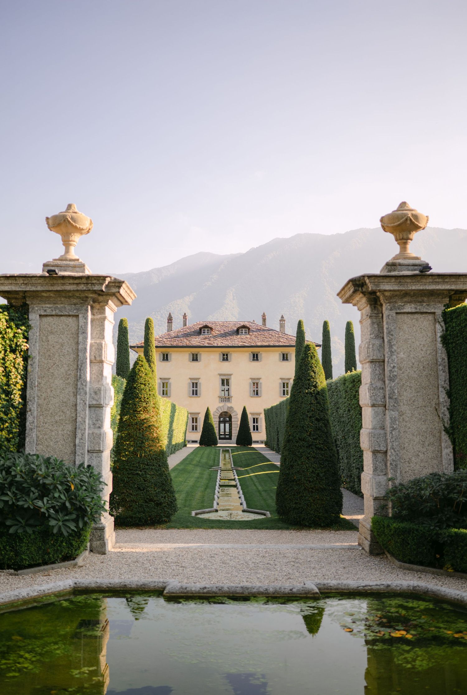 Elegant Italian villa with formal gardens, cypress trees, stone gate pillars, and mountain backdrop under a clear sky.