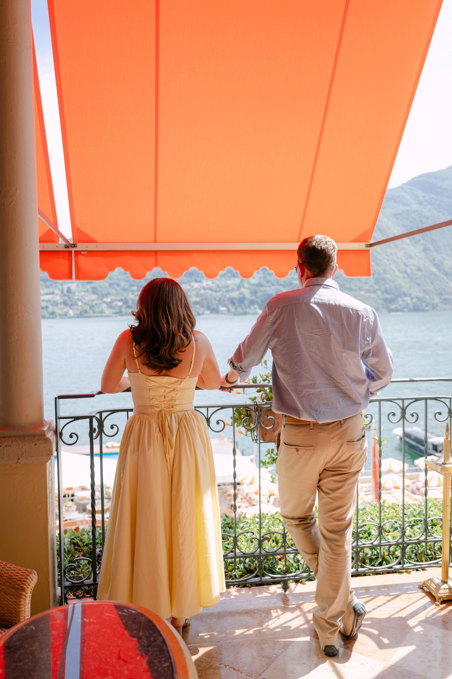 Couple standing on a scenic balcony with orange awning overlooking a lake and mountains, woman in yellow dress.