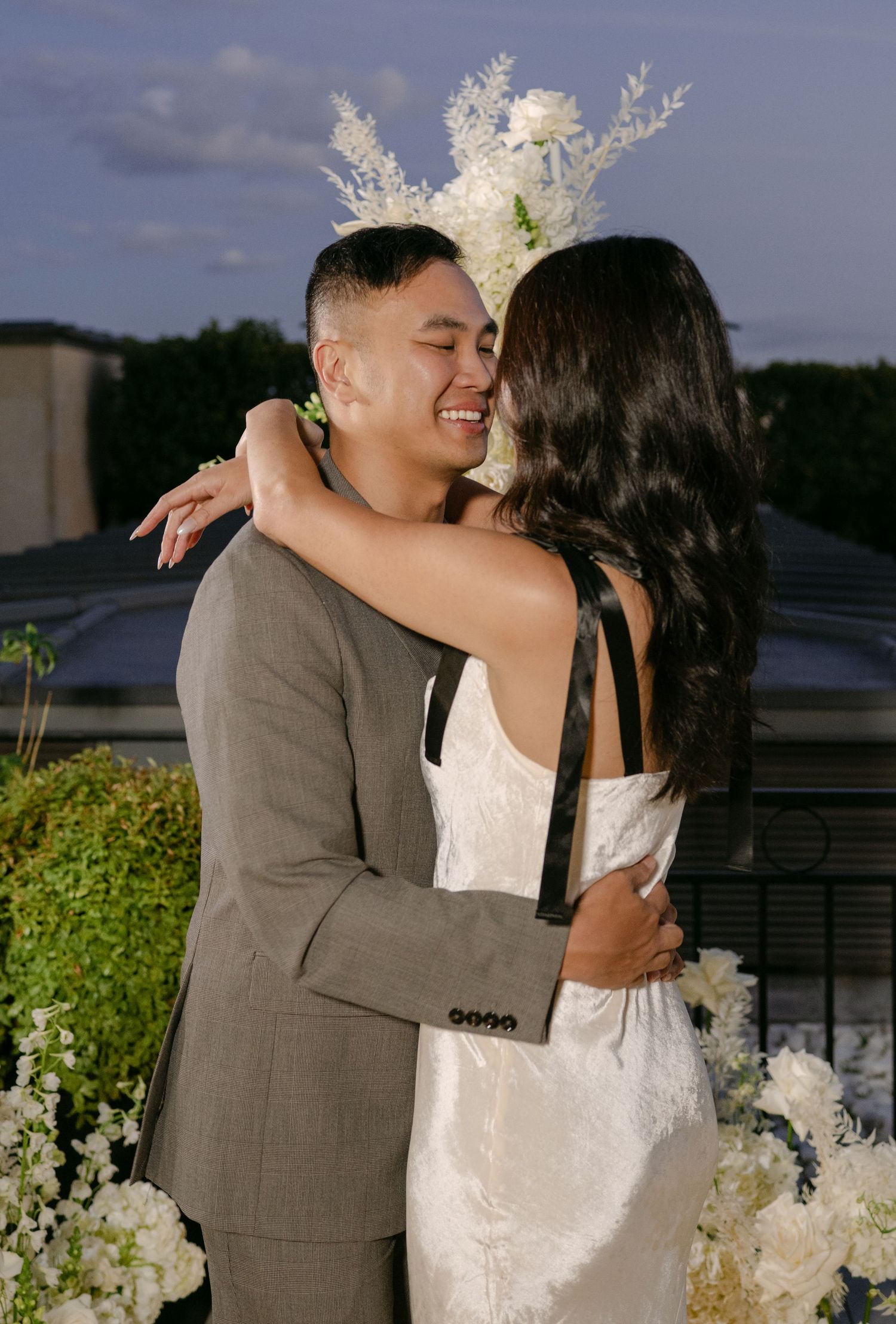 Engaged couple shares a joyful embrace beside white floral arrangements on a rooftop at dusk after a Paris proposal.