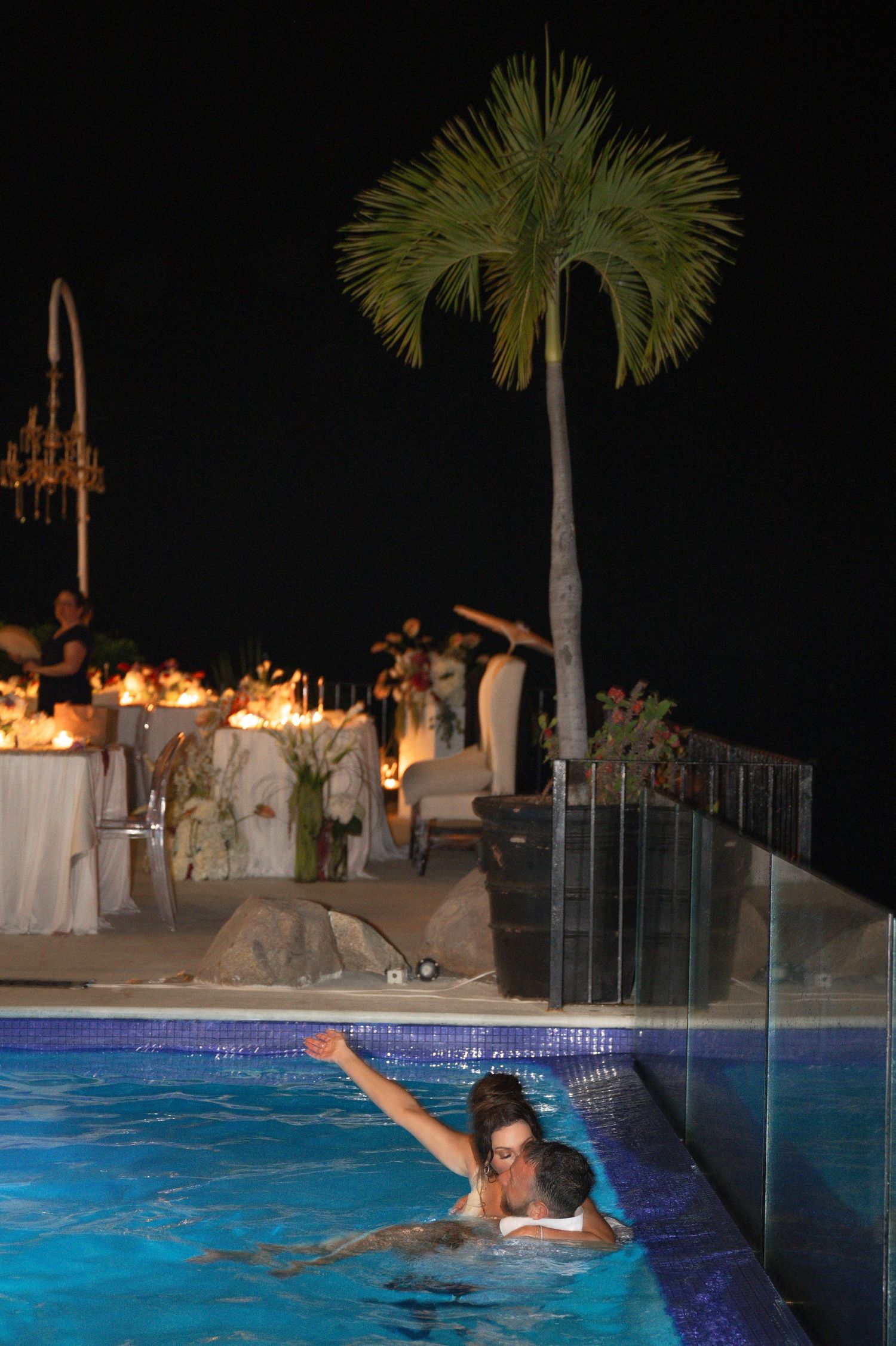 Bride and groom swimming in pool at outdoor nighttime wedding reception.