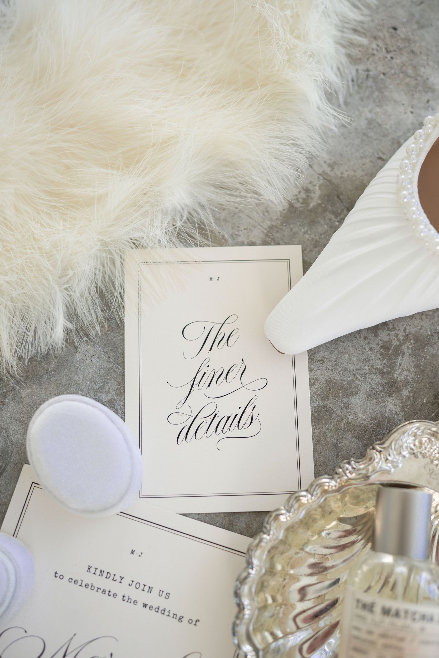 Elegant wedding flat lay with calligraphy card, white heel, fur, perfume bottle, and silver tray on marble.