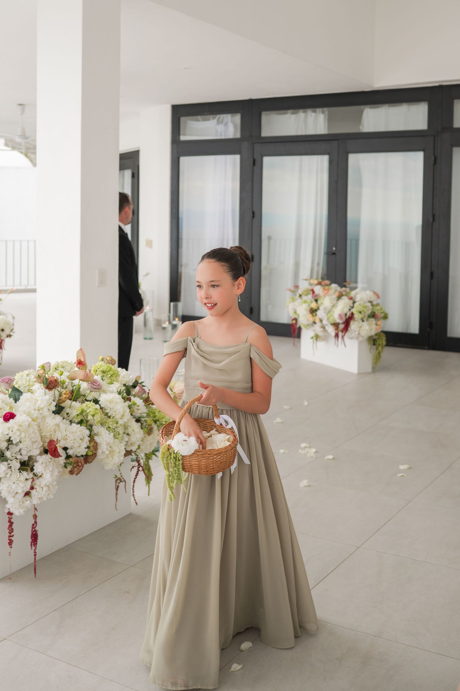 Young flower girl in sage green off-shoulder gown holds wicker basket at elegant indoor wedding ceremony.