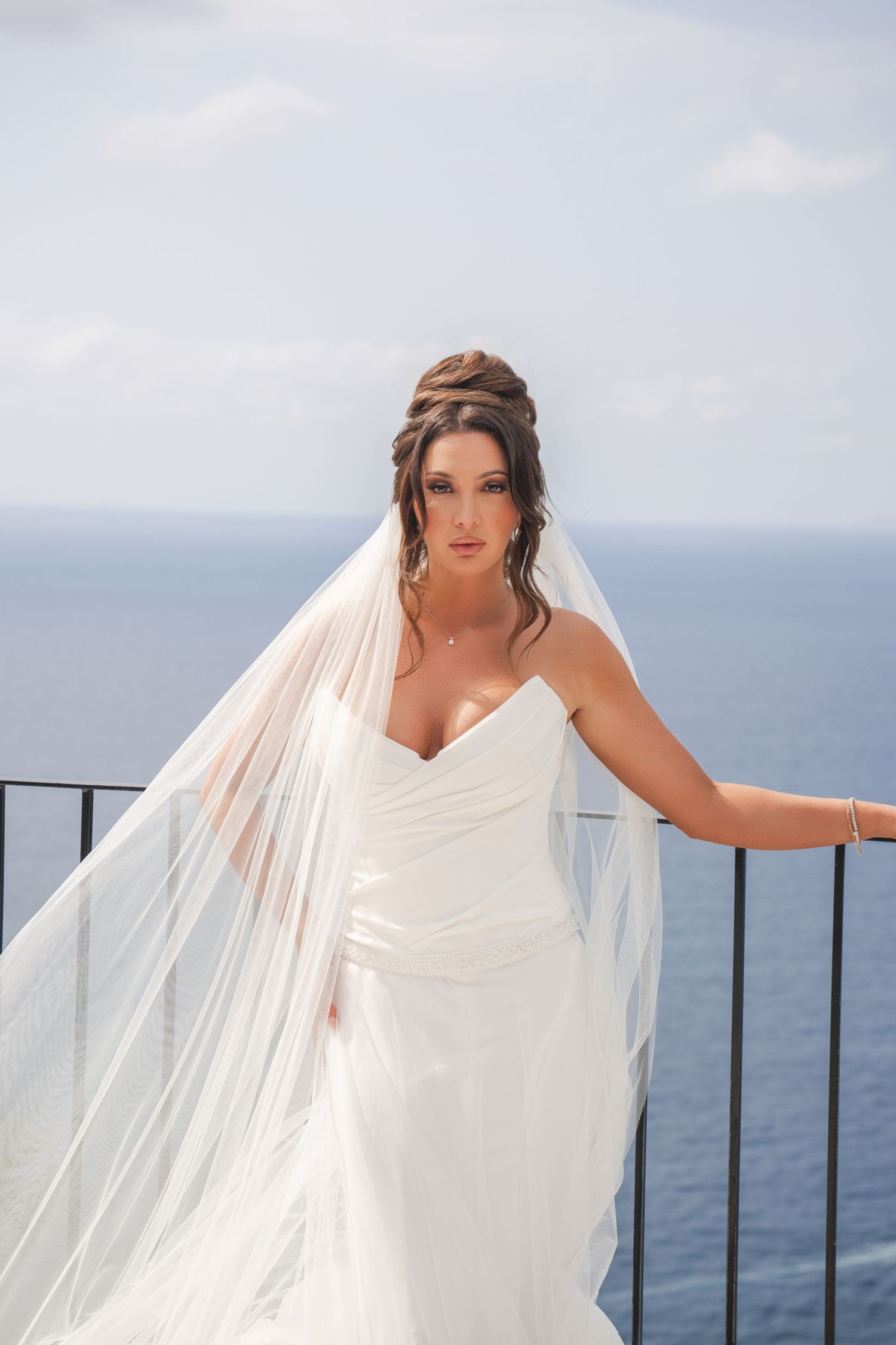 Elegant bride in white gown and veil poses on a balcony with ocean views behind her.