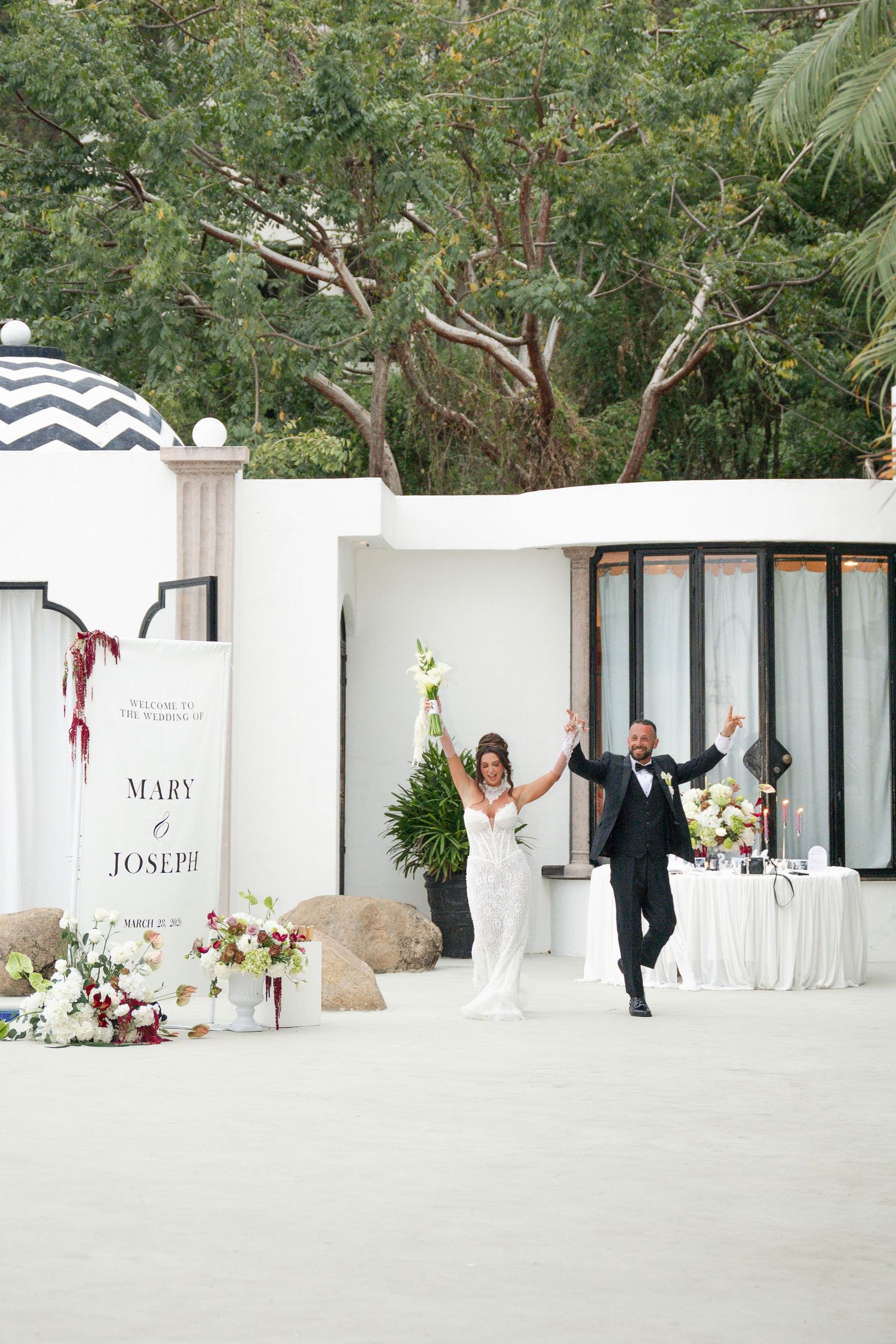 Bride and groom celebrating at outdoor wedding reception, arms raised joyfully near Mary and Joseph wedding sign.