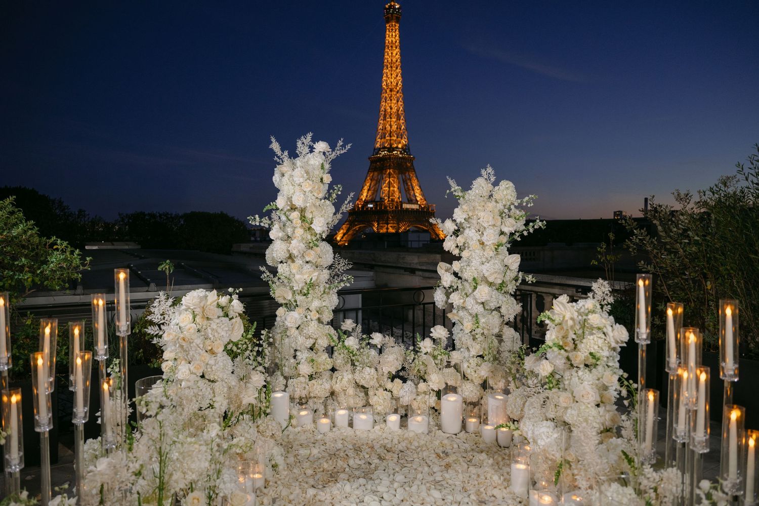 Romantic Paris wedding setup with white floral arrangements and glowing candles at dusk, overlooking the illuminated Eiffel Tower.