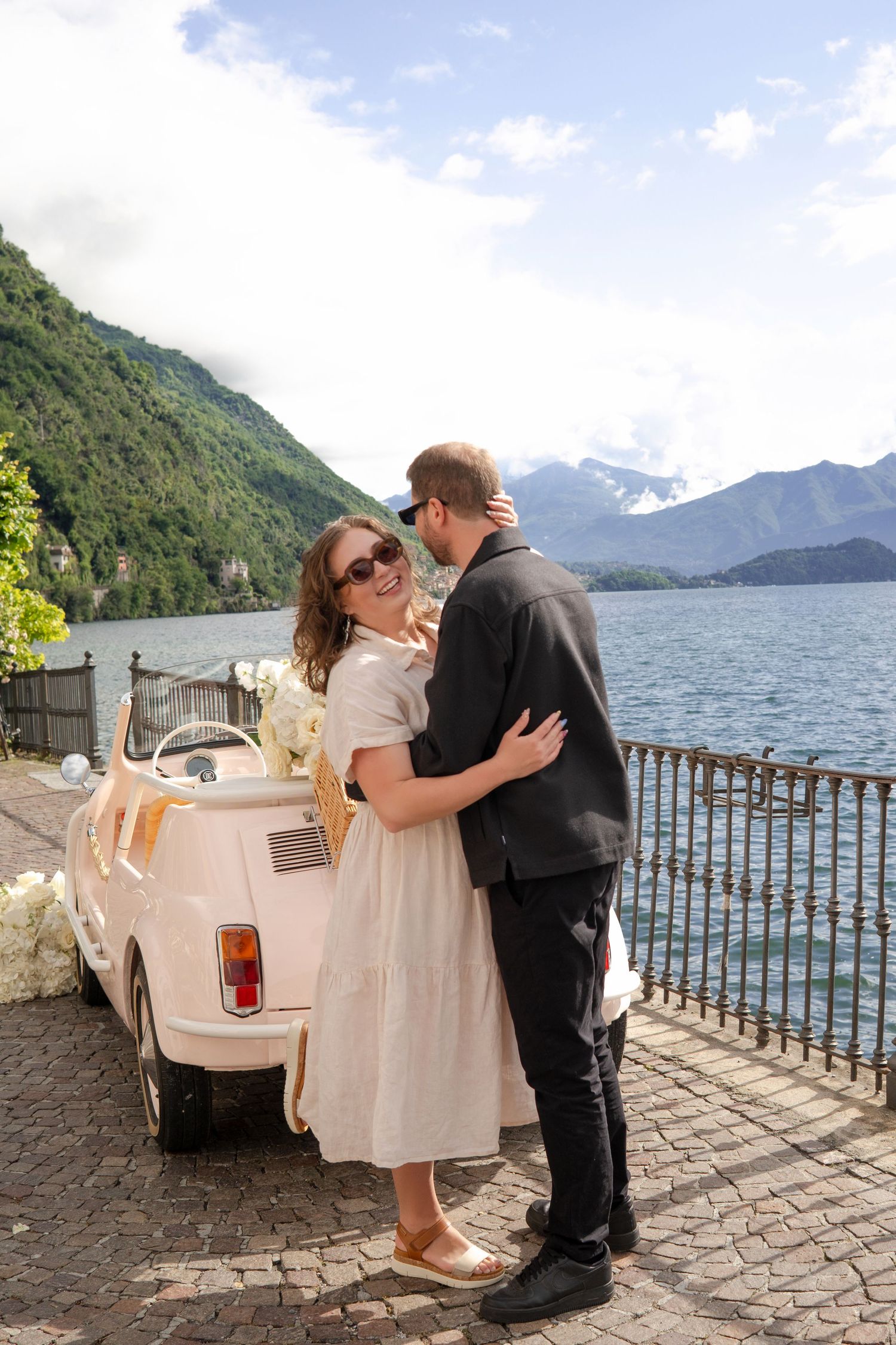 Couple embracing near a vintage pink Fiat 500 by Lake Como, Italy, with mountain backdrop on a sunny wedding day.