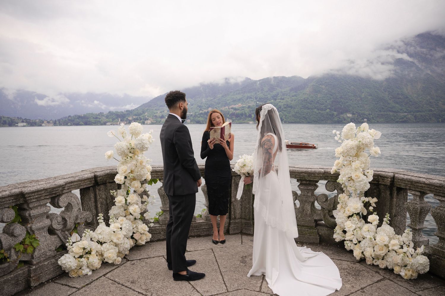 Couple exchanges vows on a lakeside terrace with white floral arrangements and misty mountains in the background.