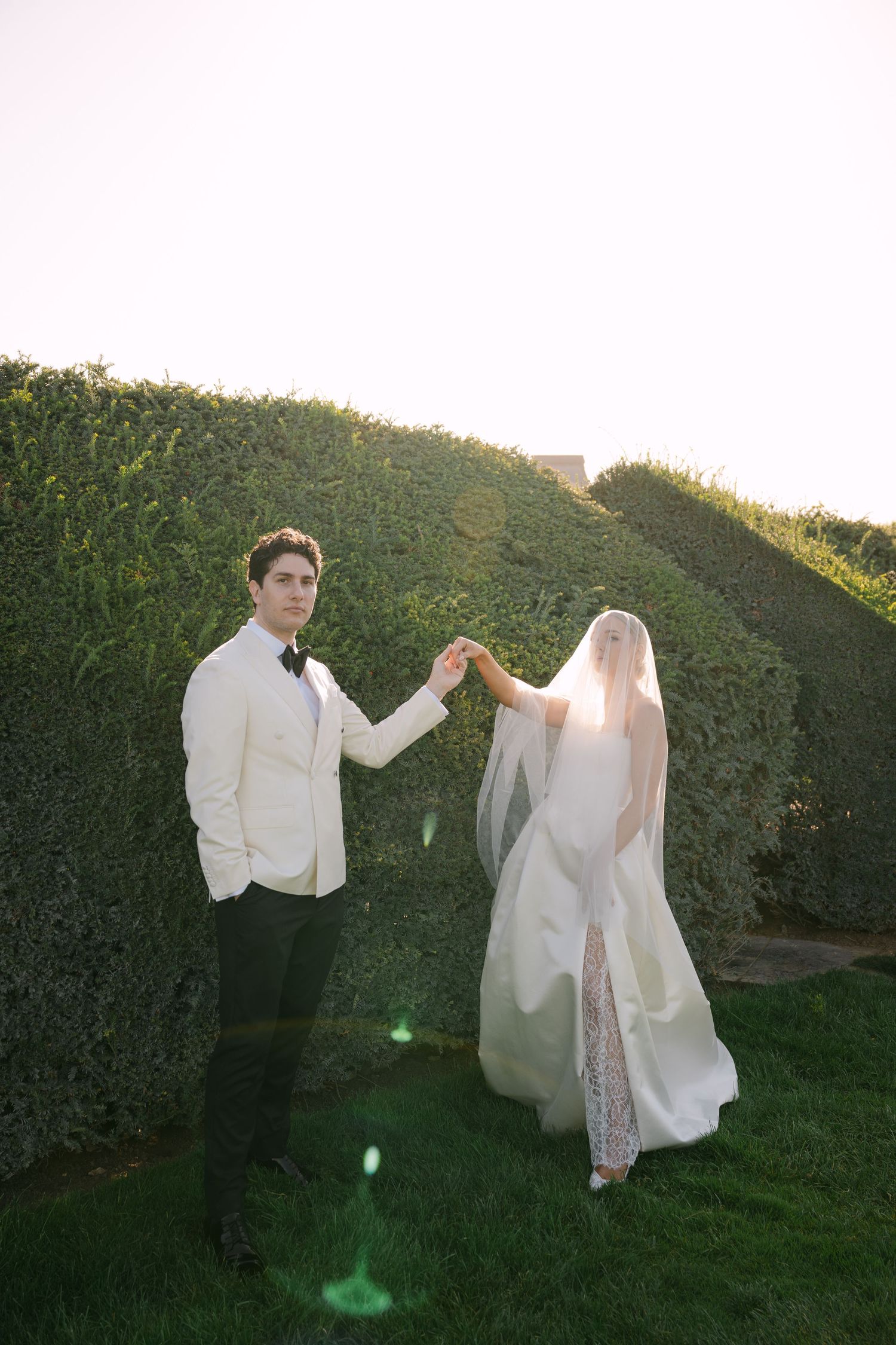 Bride and groom hold hands in a lush green hedge garden, groom in white tuxedo, bride in flowing white gown with veil.
