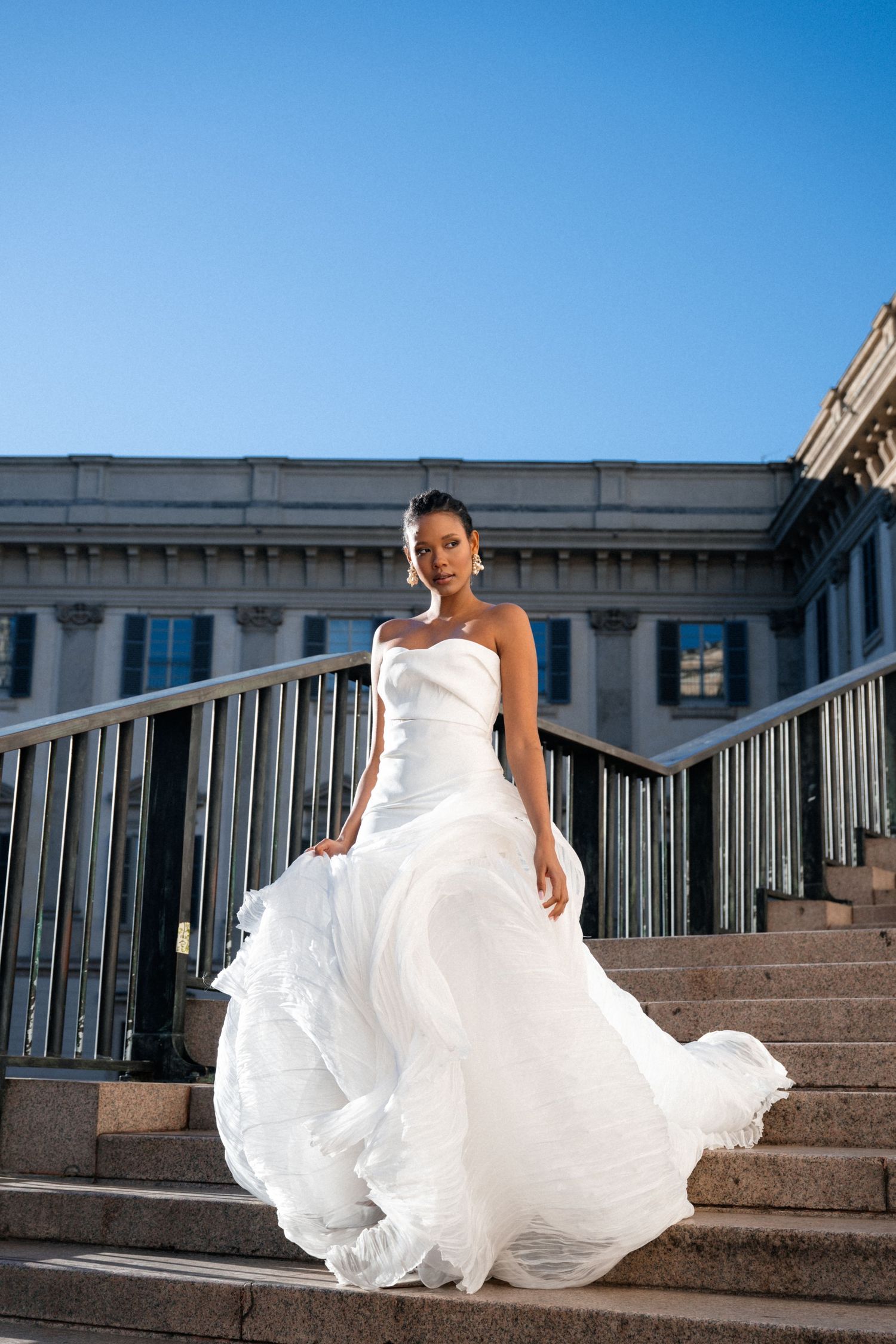 A Black bride in a flowing white strapless ball gown stands on stone steps outside a classical building.