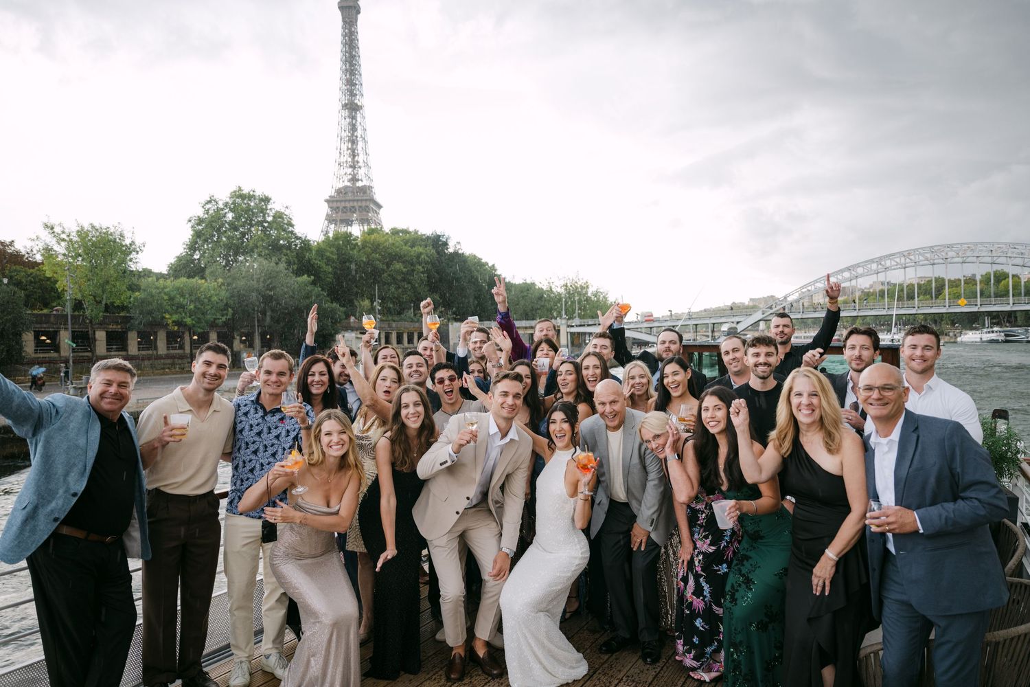 Large wedding party gathers on a deck overlooking water and cityscape at twilight.