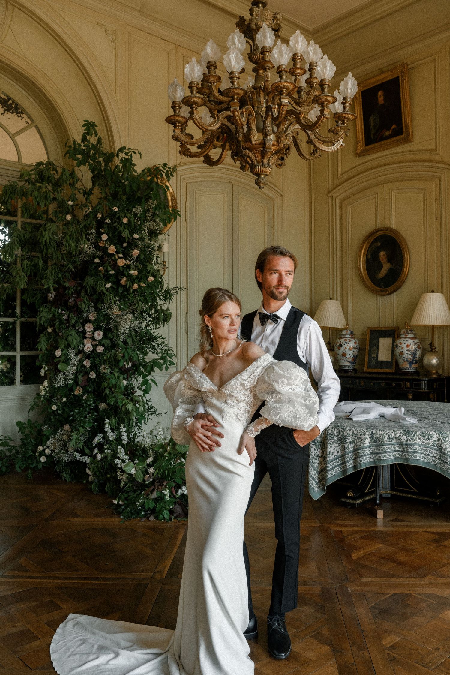 An elegant wedding couple poses in a luxurious room with a crystal chandelier and floral arrangements.