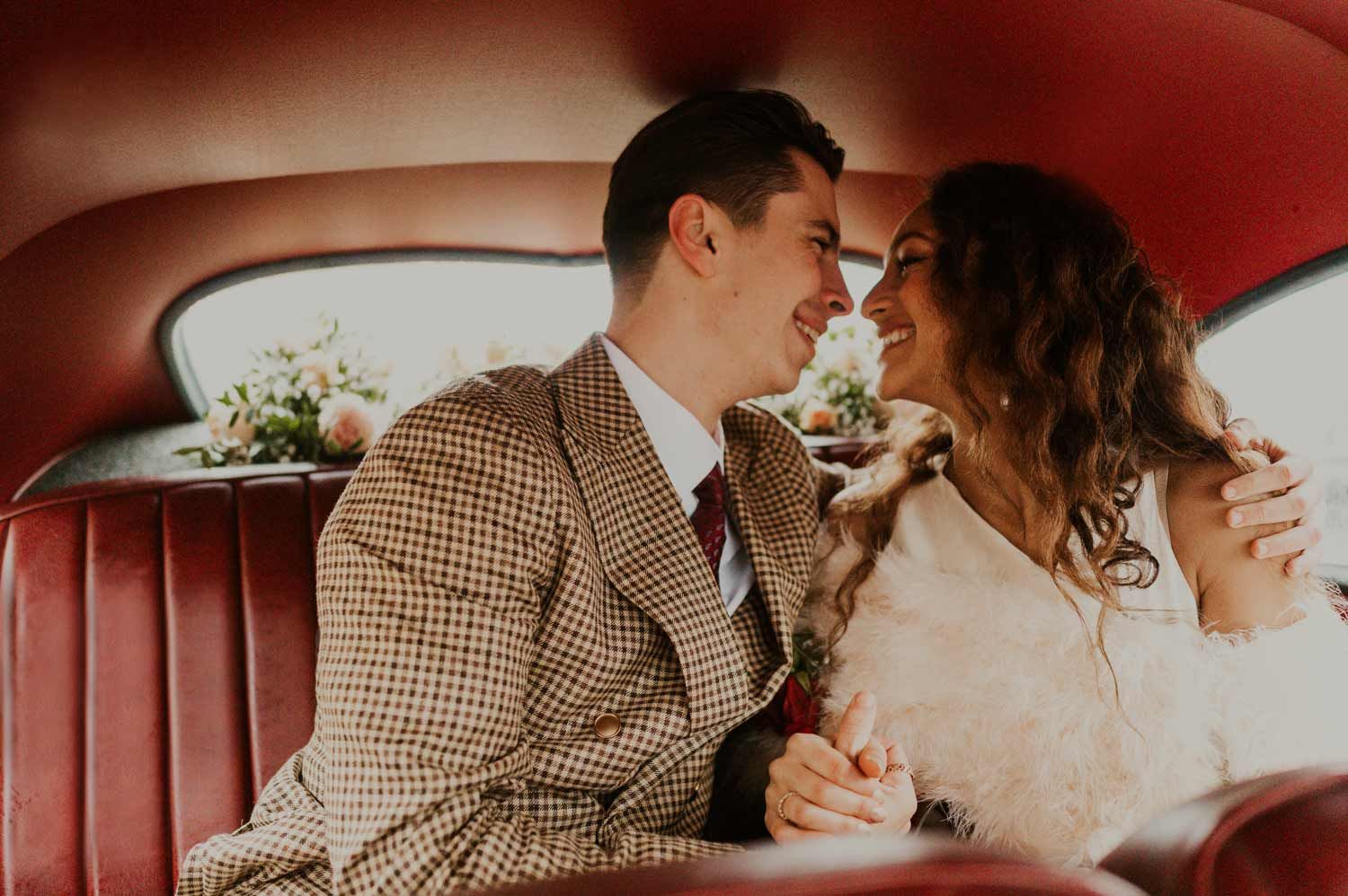 Newlyweds share a joyful moment in the back of a classic car with red leather seats.