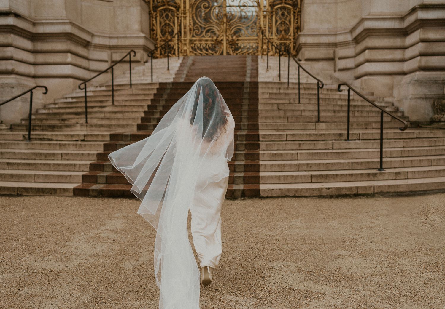 A bride in a flowing white gown stands on stone steps with elegant architecture in the background.