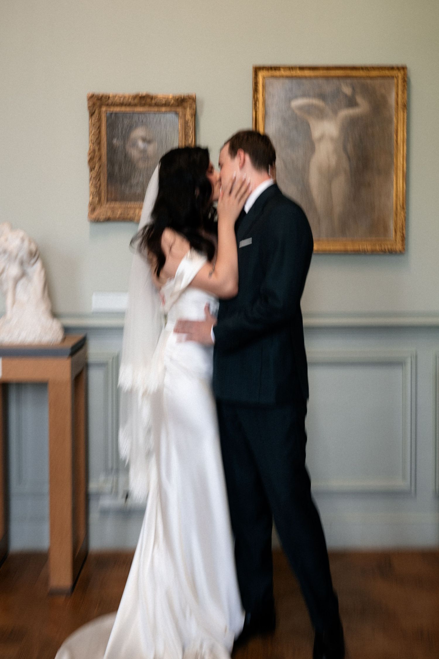 Bride and groom share a first kiss surrounded by classical paintings and sculptures in a beautiful art museum.