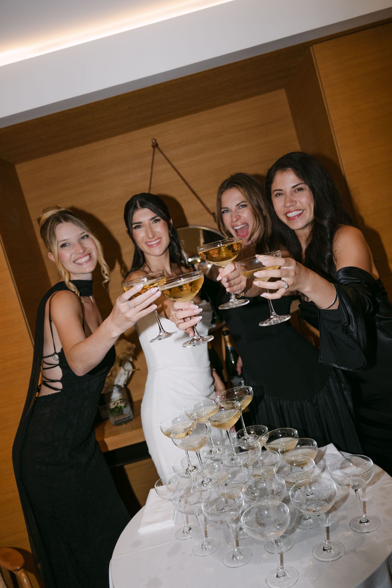 Bride and bridesmaids in black dresses toasting champagne glasses at wedding reception.