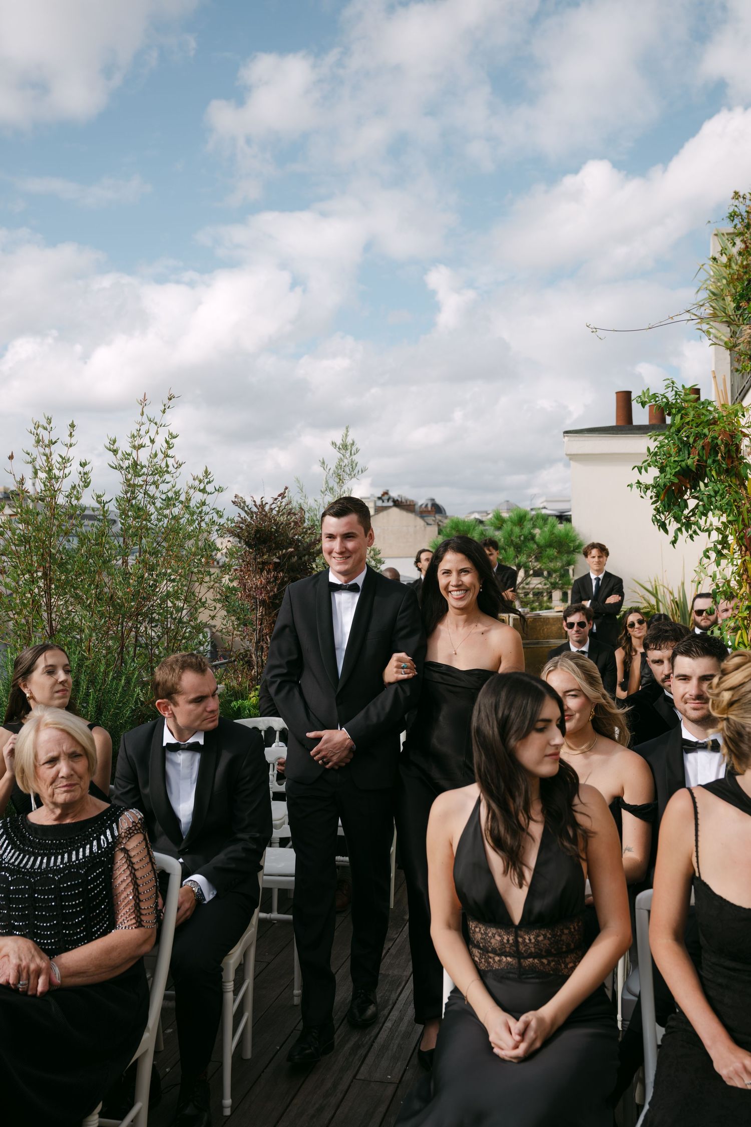 Groomsman escorts woman in black dress down outdoor wedding aisle with seated guests on both sides.