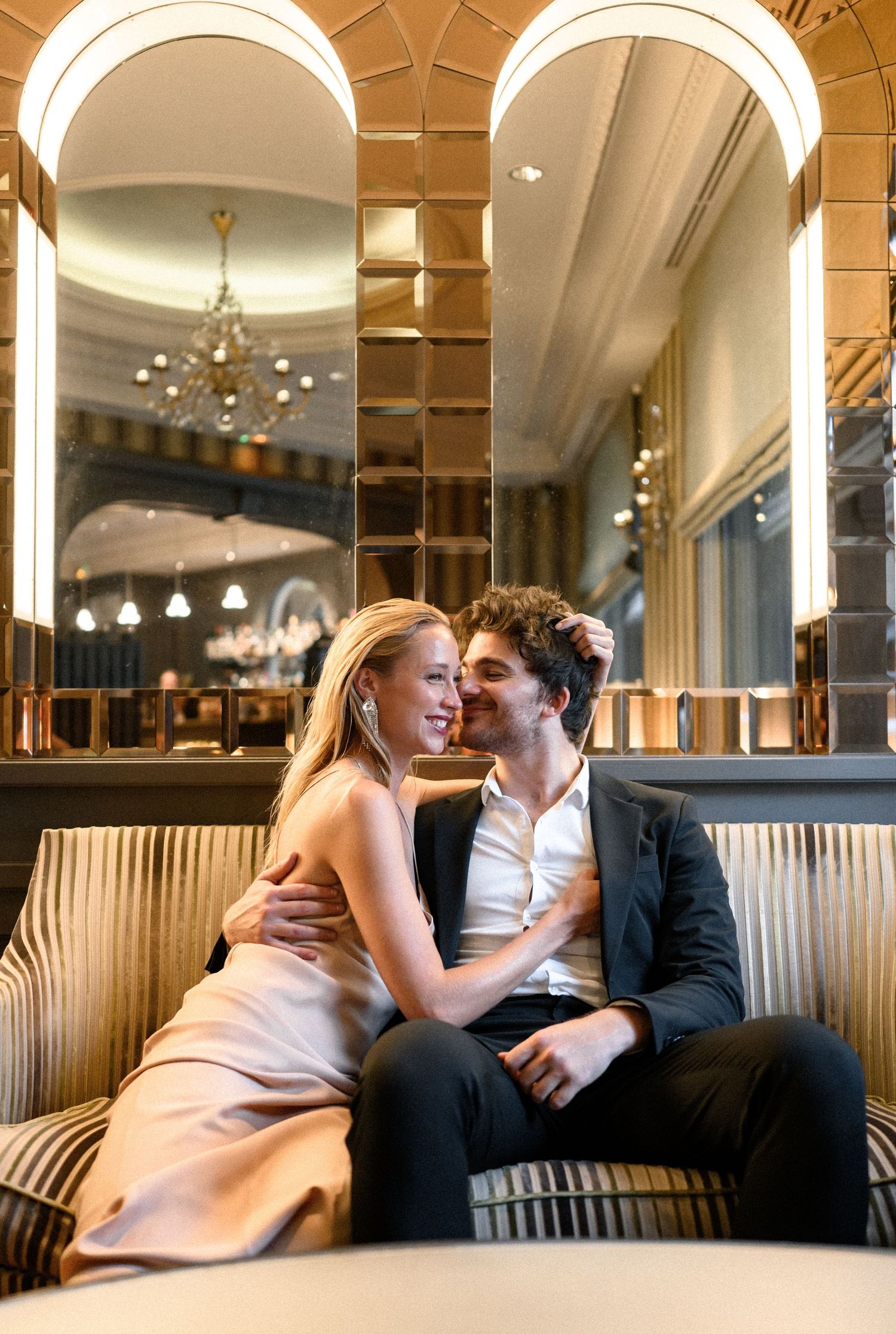 Couple sharing a tender kiss while seated in an elegant hotel lounge with arched mirrors.