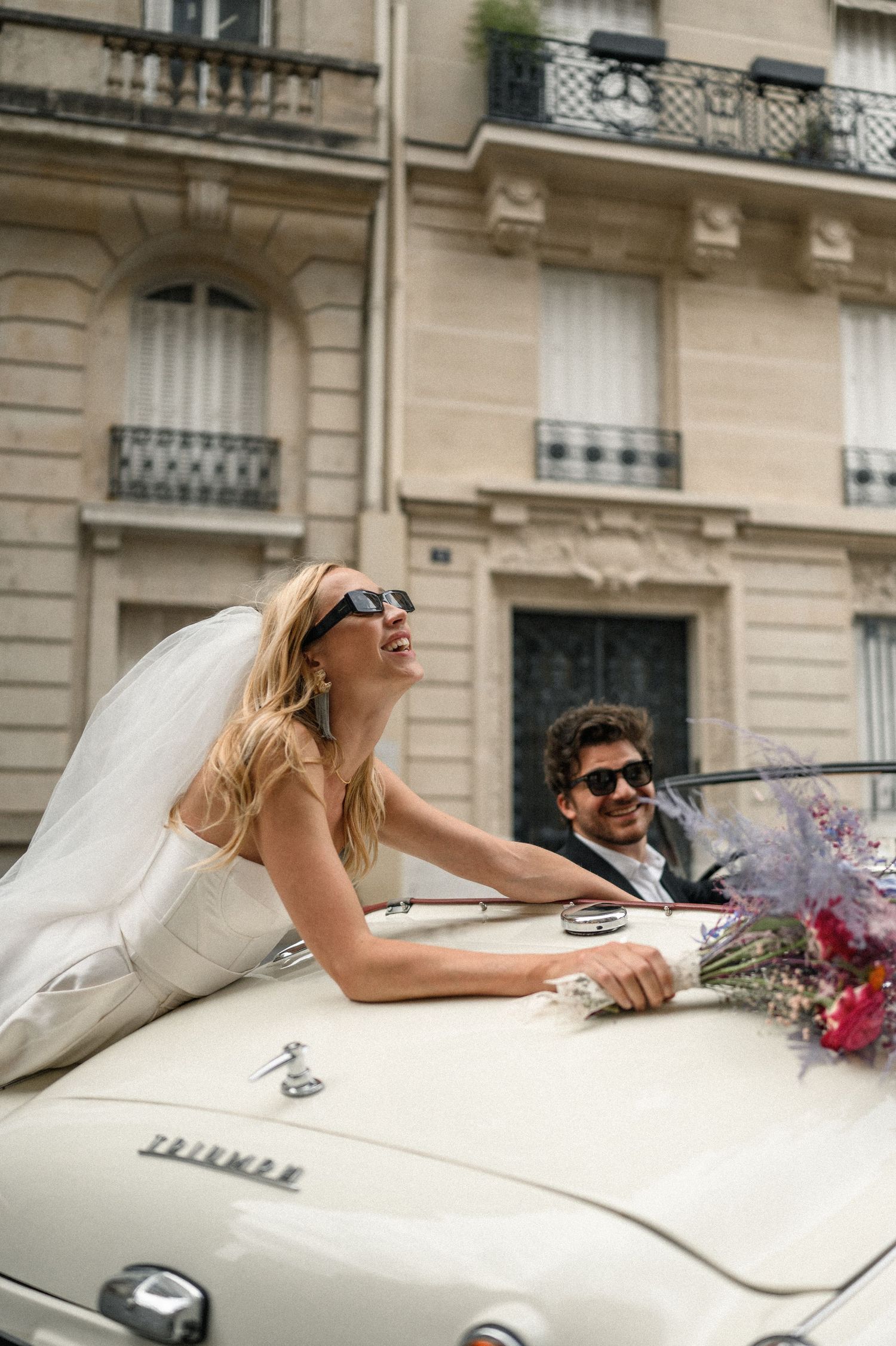 Laughing bride leans out of vintage white Triumph convertible with groom driving through elegant Paris neighborhood.