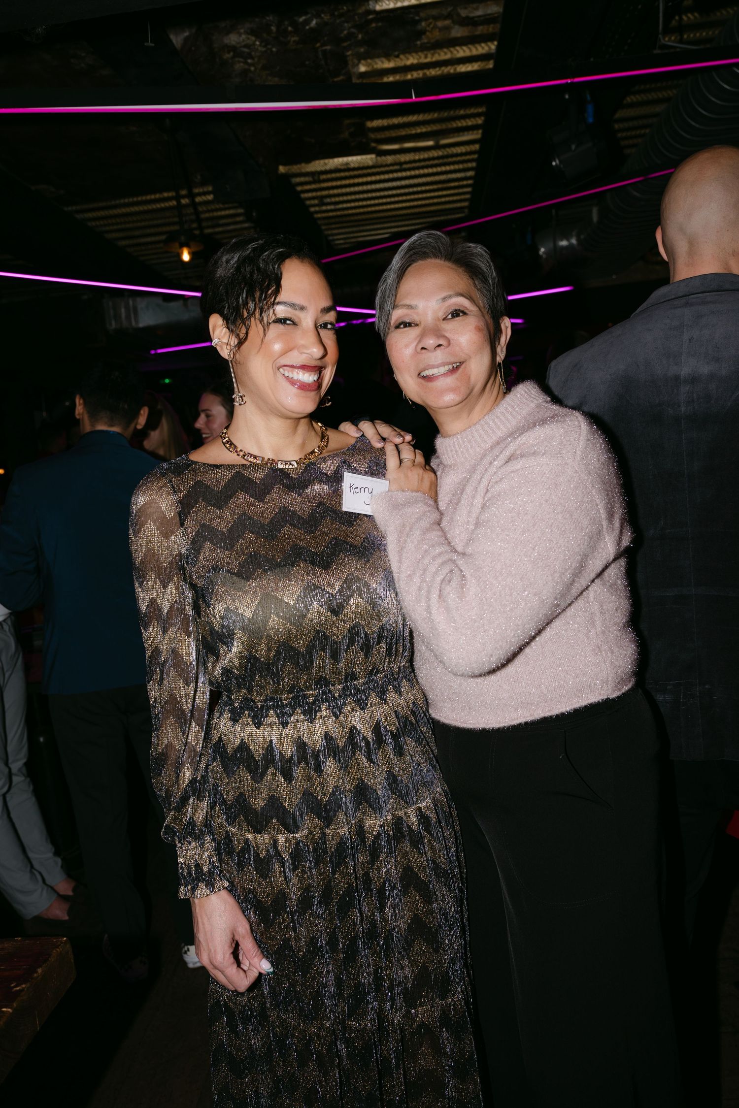 Two women smiling and posing together at an evening event, one in a gold zigzag dress and one in a beige sweater.