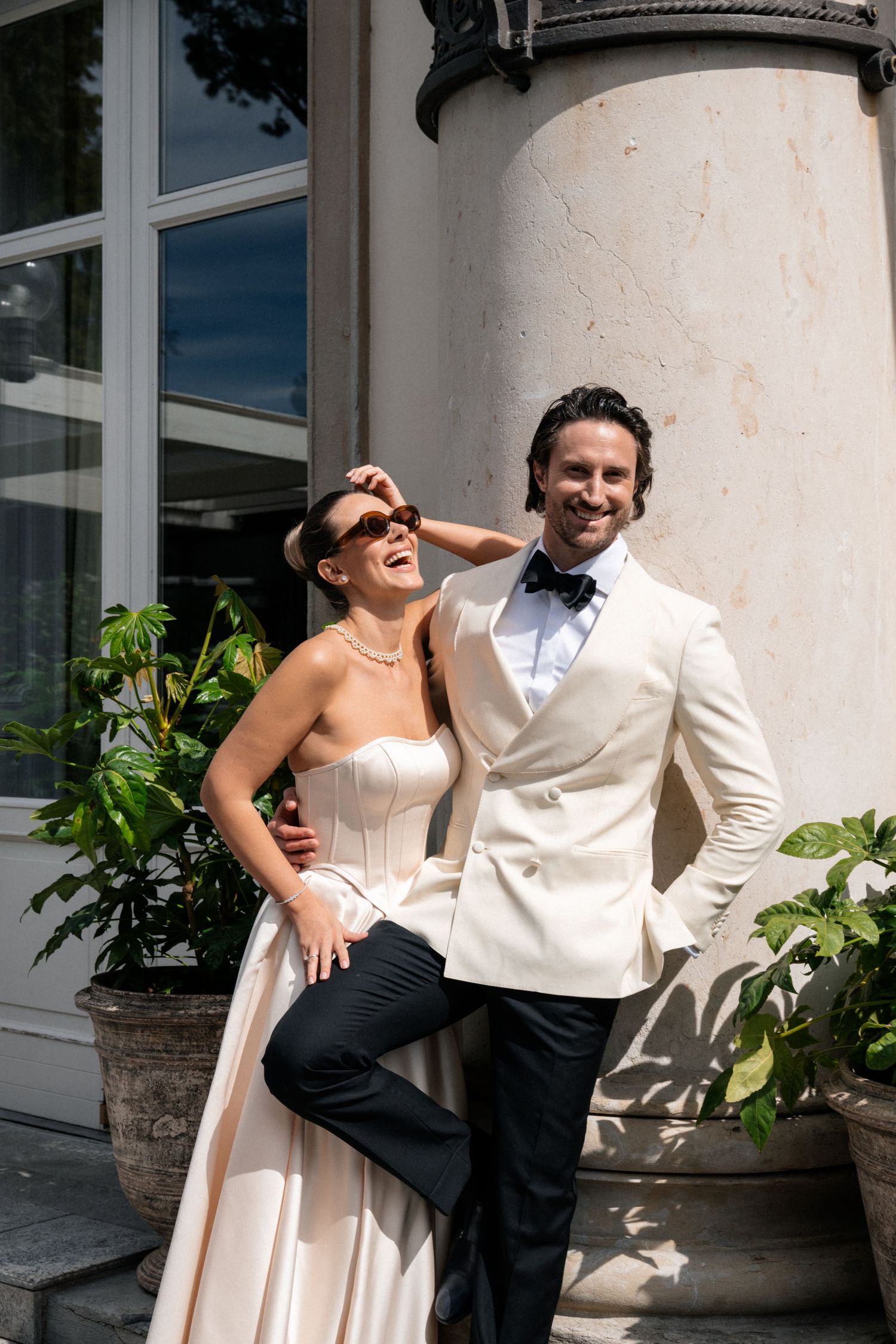 Stylish wedding couple in champagne and ivory attire laugh together beside classical stone columns.