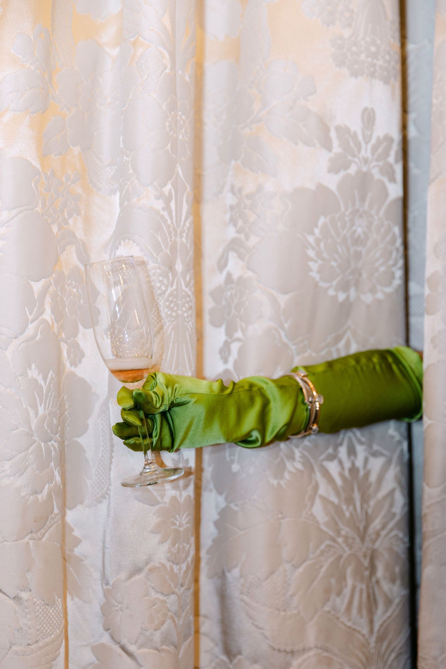 An elegant arm in a green satin glove with a gold bracelet holds a wine glass against ornate white damask curtains.