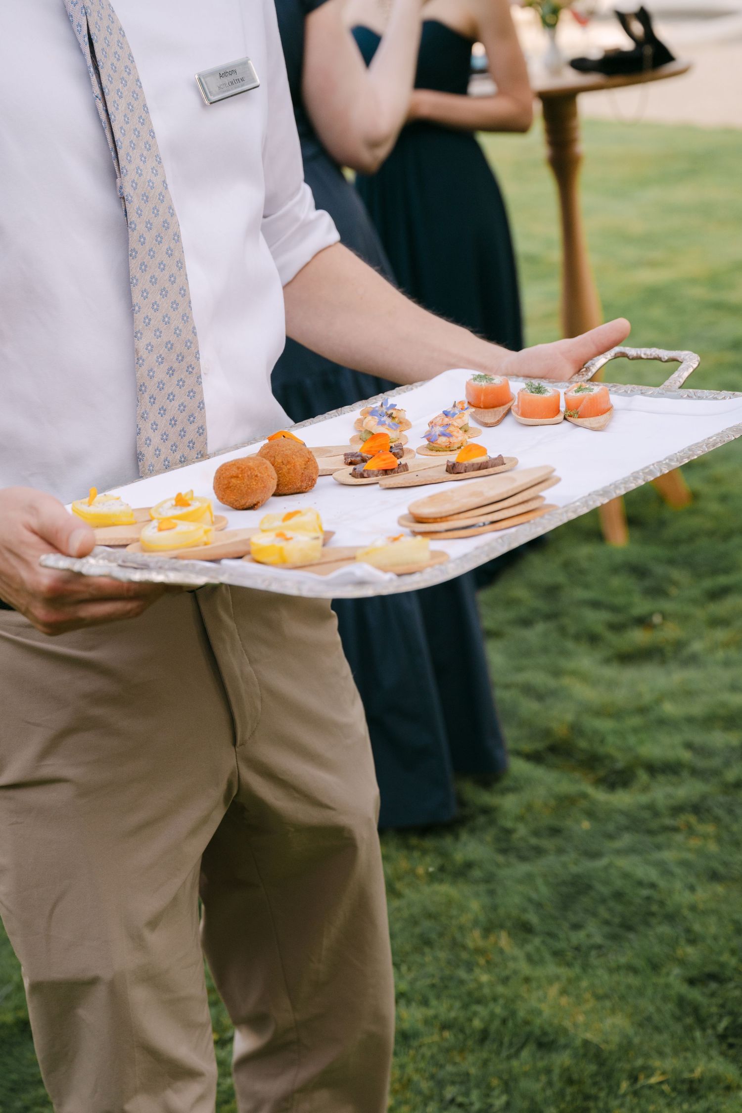 Waiter in formal attire presenting a tray of elegant hors d'oeuvres including persimmons and crackers outdoors.
