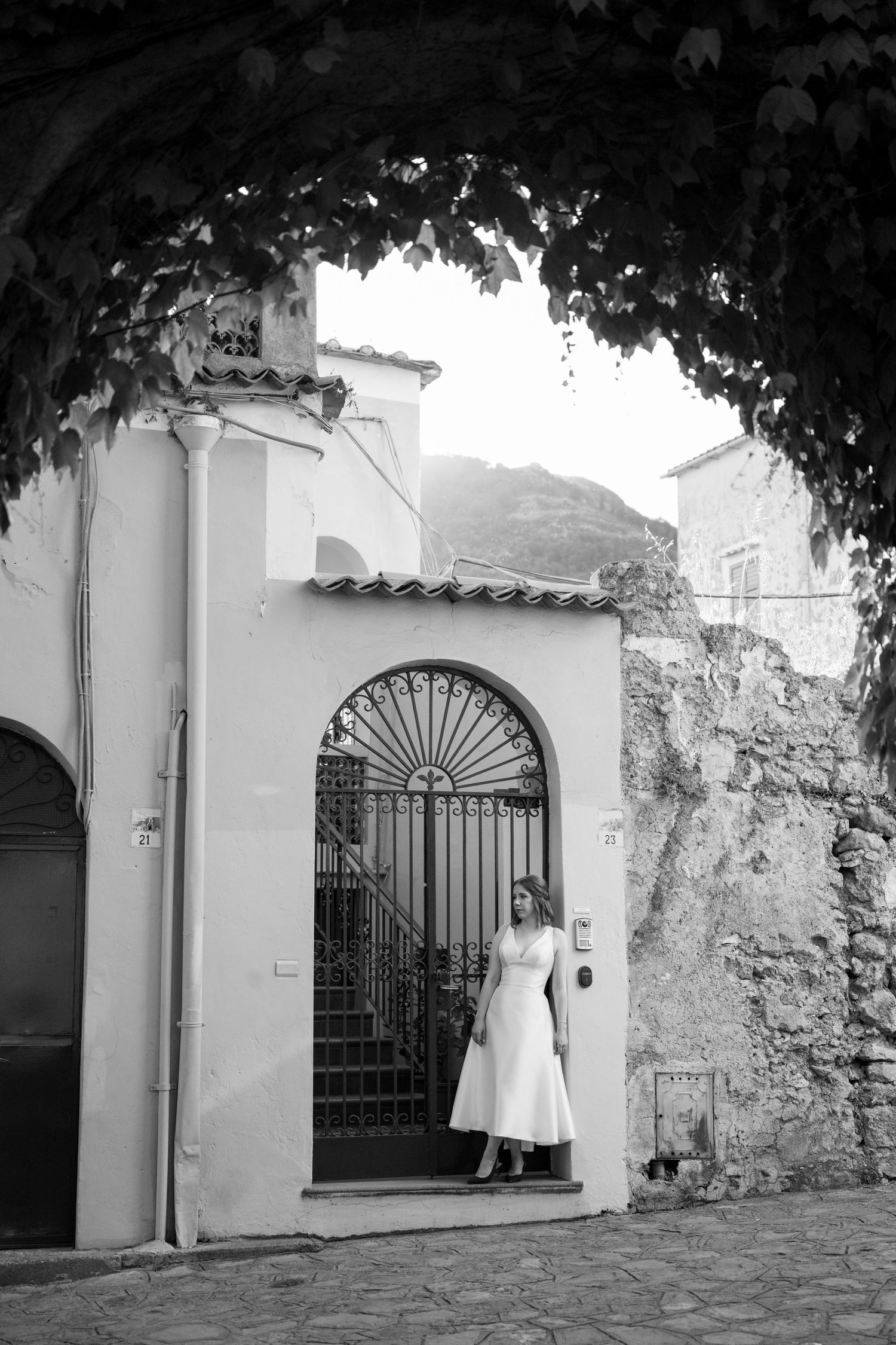 Bride in white midi dress stands in arched iron gate doorway in black and white Italian village photo.