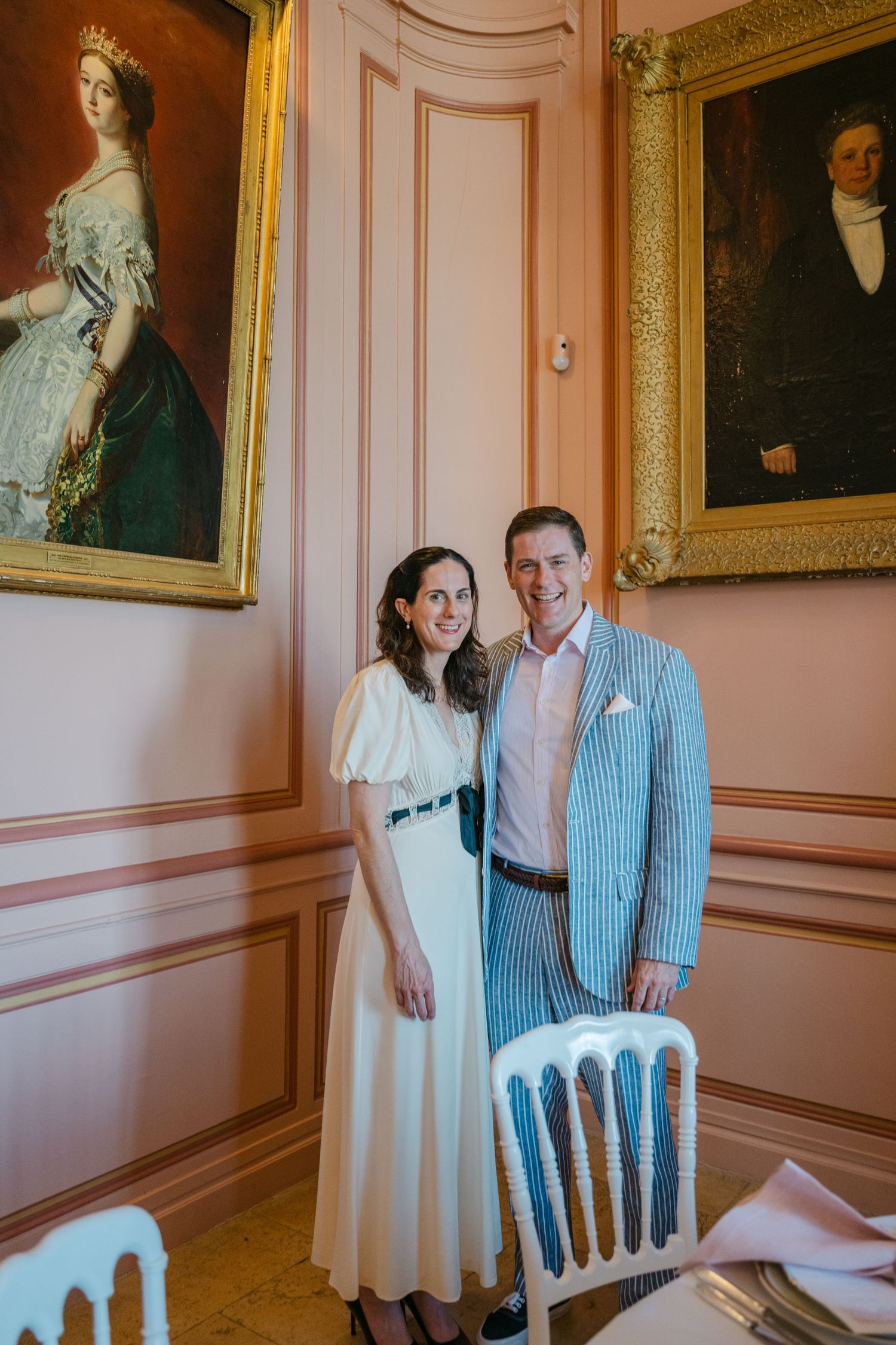 Couple smiling together in a French chateau dining room, man wearing a blue seersucker suit beside vintage portraits.