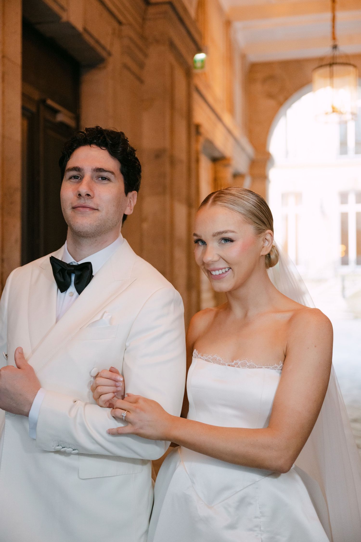 Bride and groom pose elegantly in a Parisian archway, groom in white tuxedo with black bow tie, bride in strapless gown.
