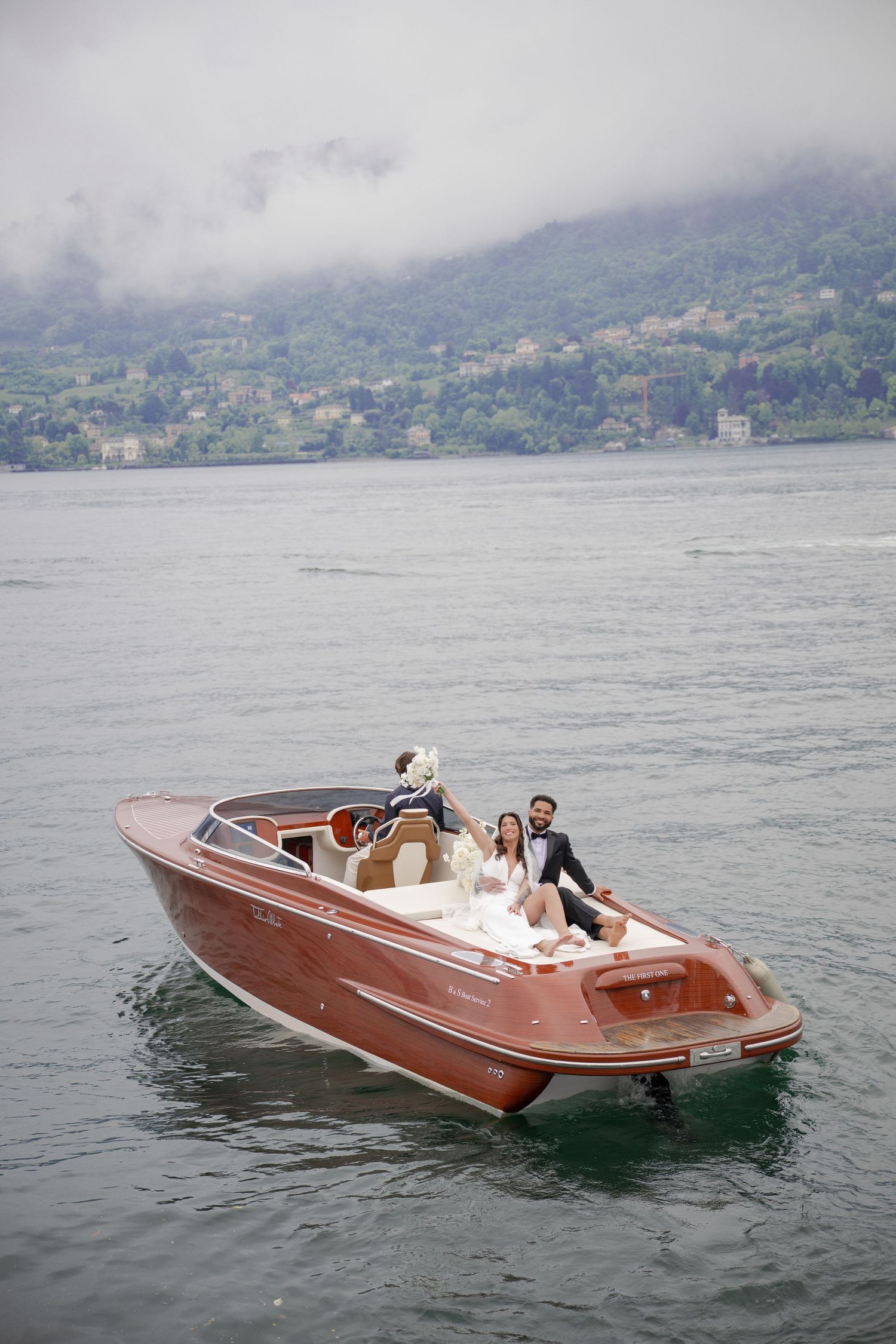 Bride and groom posing on a classic wooden speedboat on misty Lake Como with mountain backdrop.