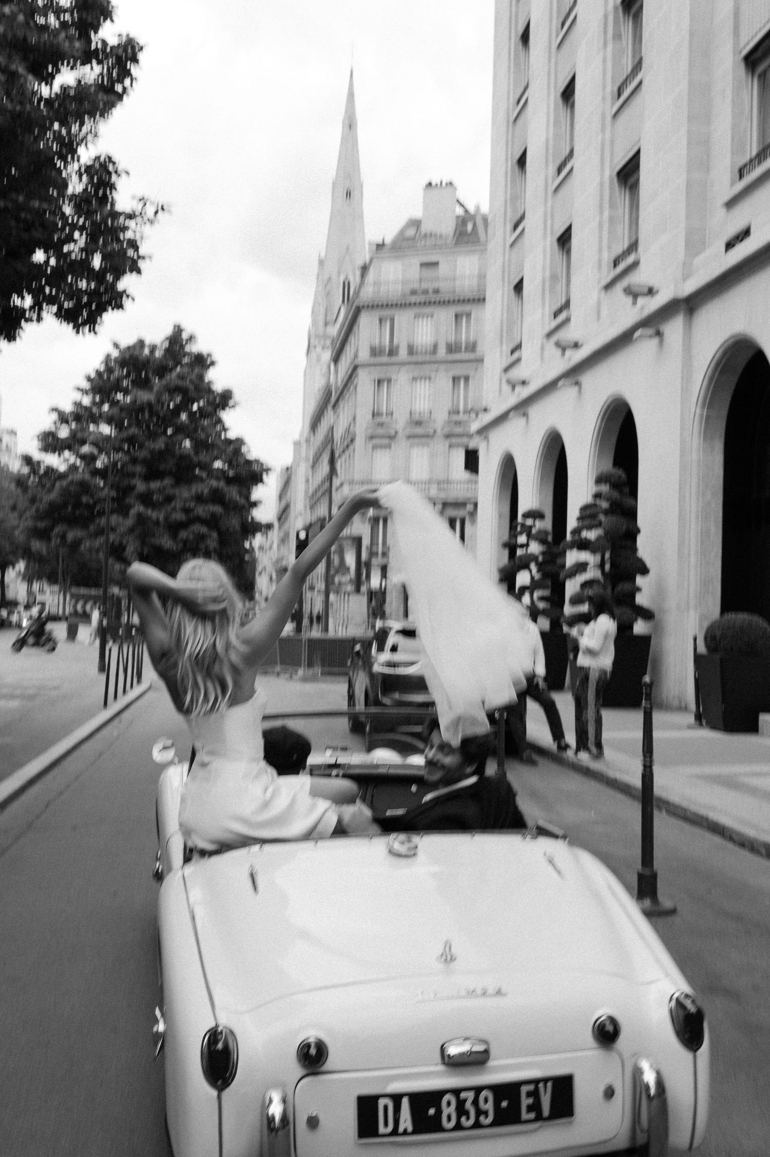 Bride in white dress waves from classic convertible car on a Parisian street in black and white photo.
