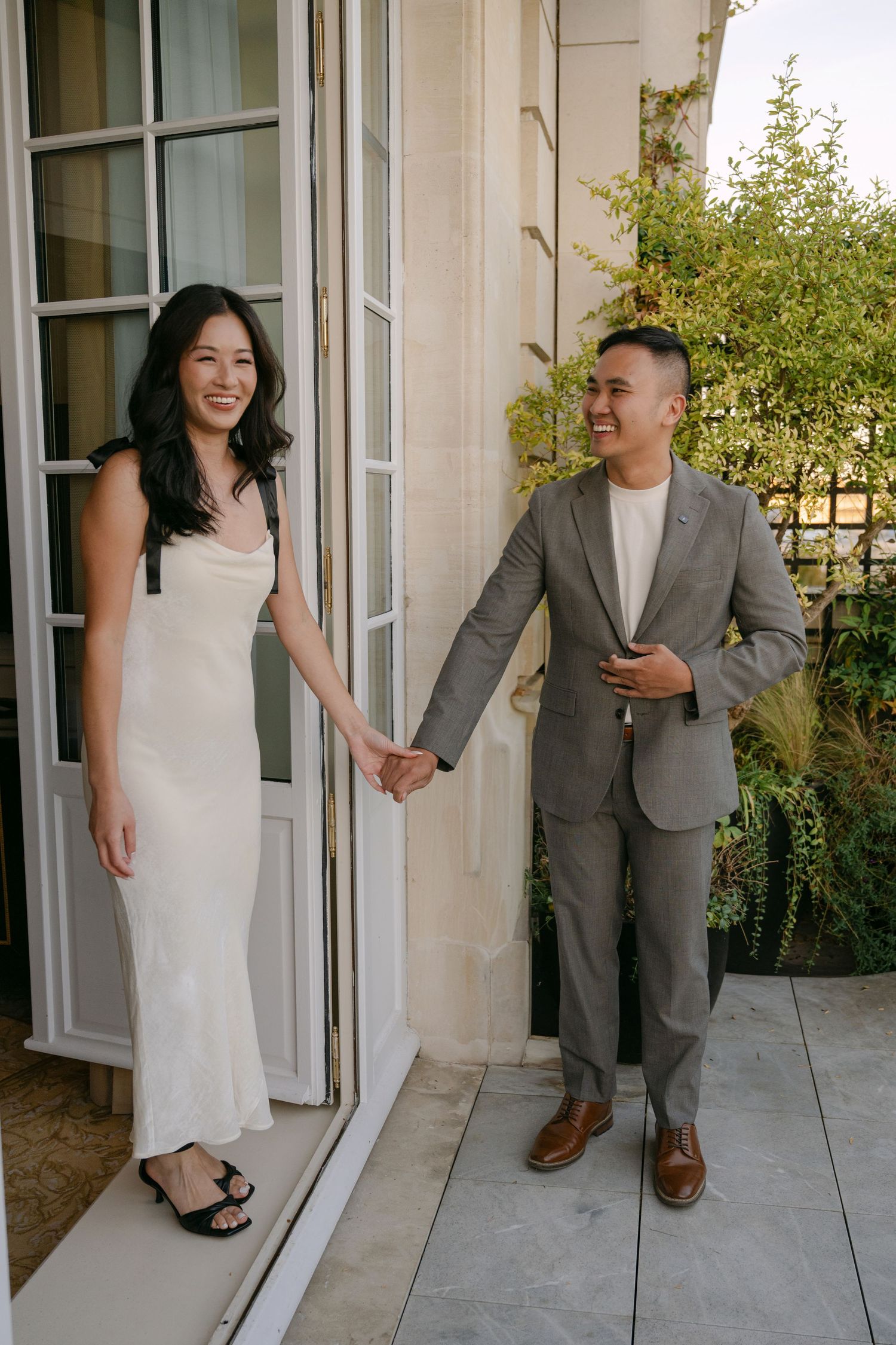Smiling couple holding hands on a sunlit terrace, she in a cream slip dress and he in a gray suit, beside French doors.