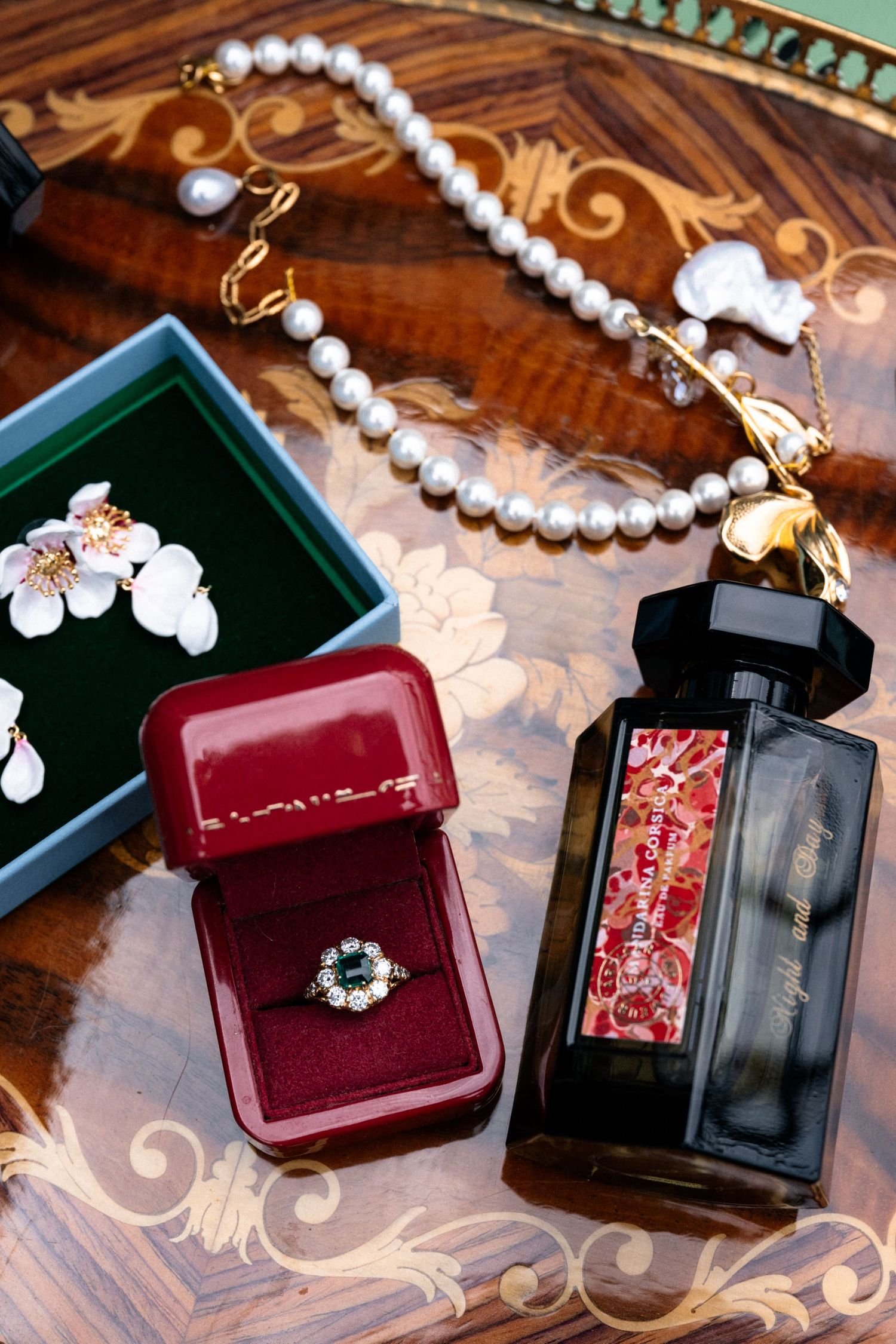 Elegant bridal flat lay with pearl necklace, emerald ring in red box, floral earrings, and perfume bottle on ornate wooden table.