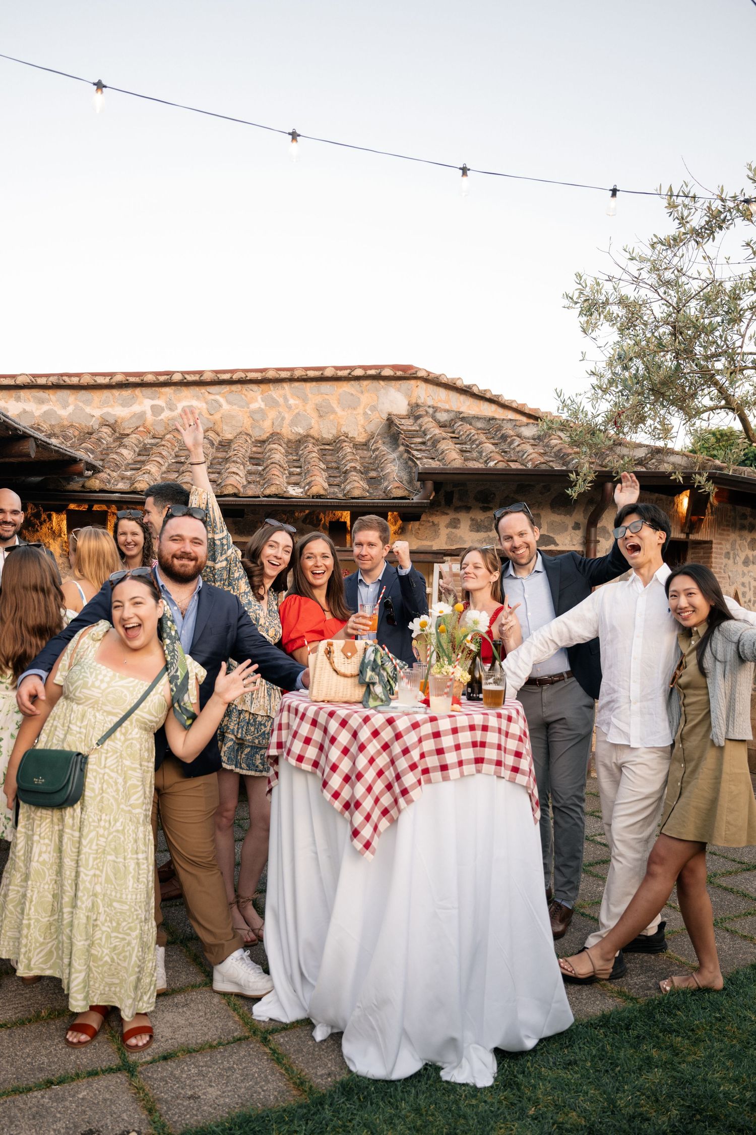 Wedding party group photo with bride and groom at red checkered table under string lights outdoors.