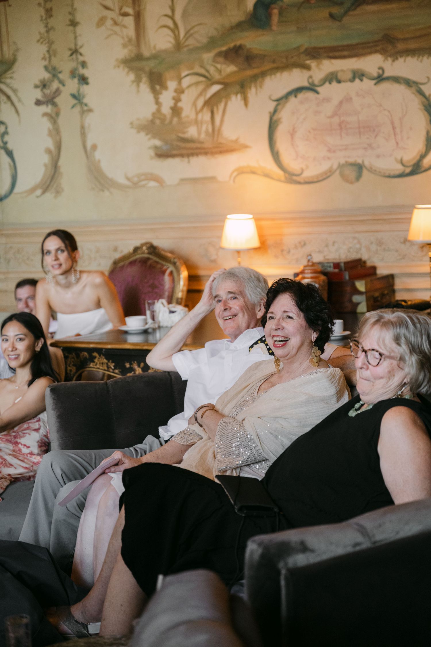 Wedding guests laugh and smile while seated on sofas in a beautifully decorated French chateau room.