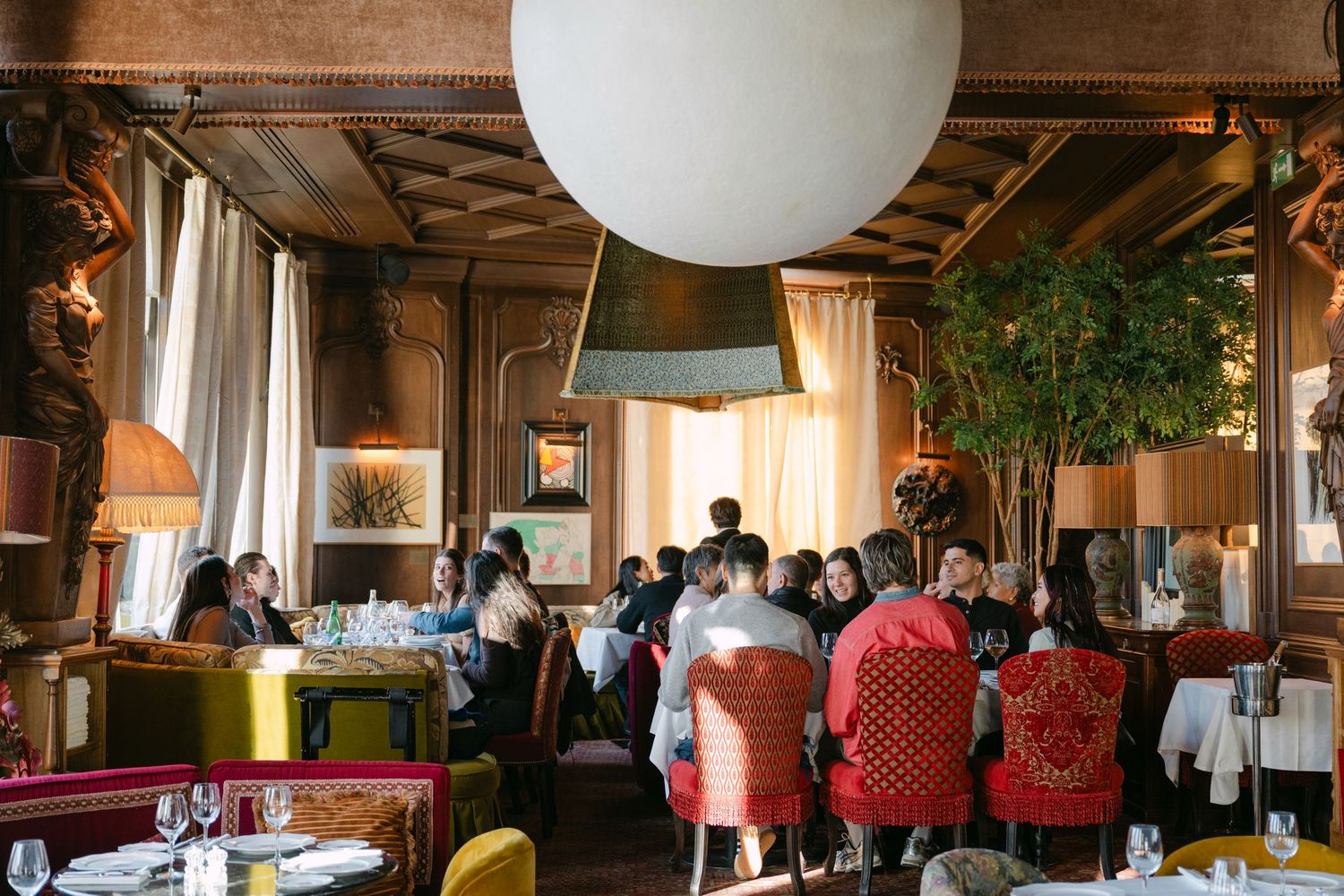 Guests dining in an upscale restaurant with eclectic decor, lush indoor trees, and warm ambient lighting.