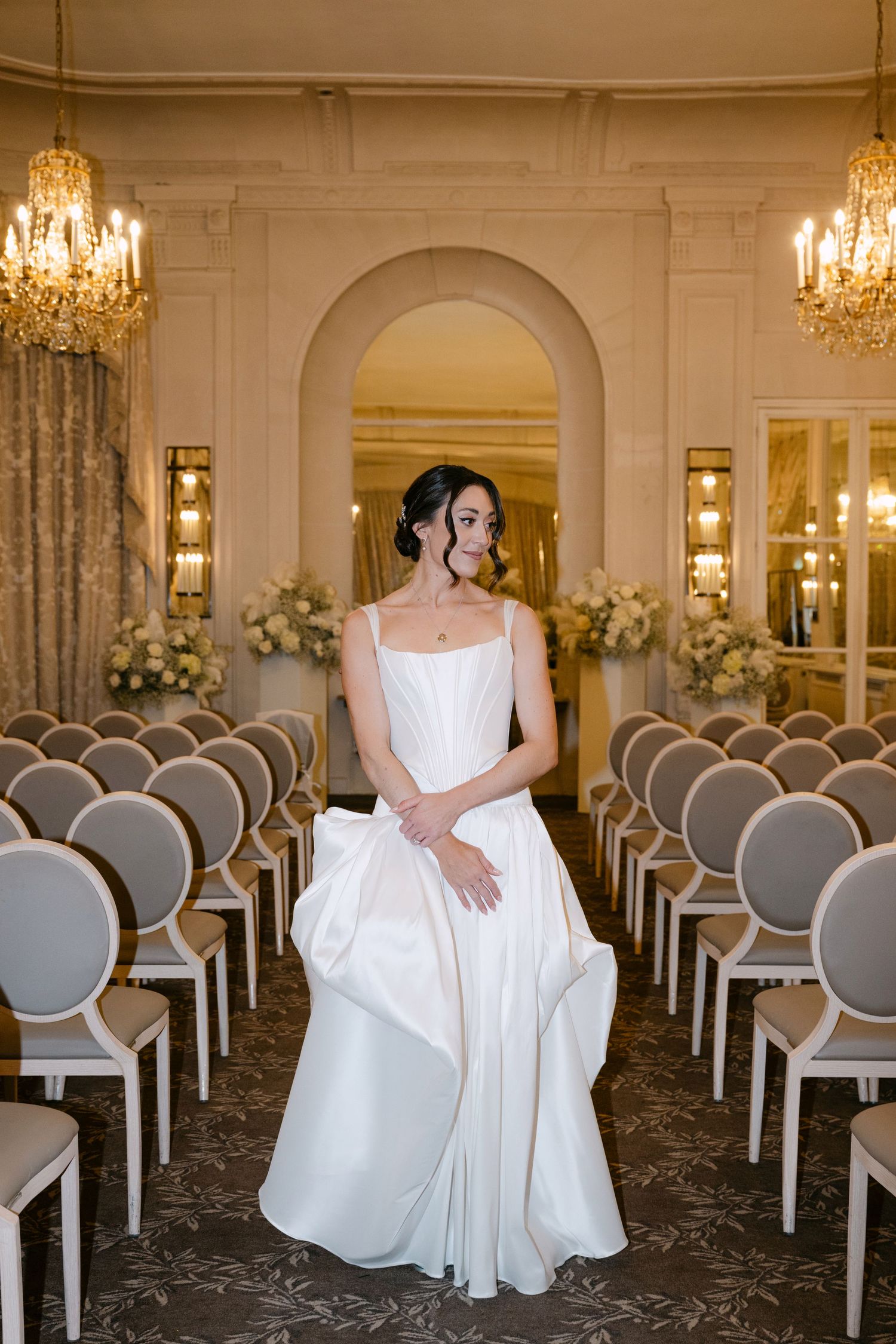 Bride in white satin A-line gown standing in elegant chandelier-lit ceremony room with rows of chairs.